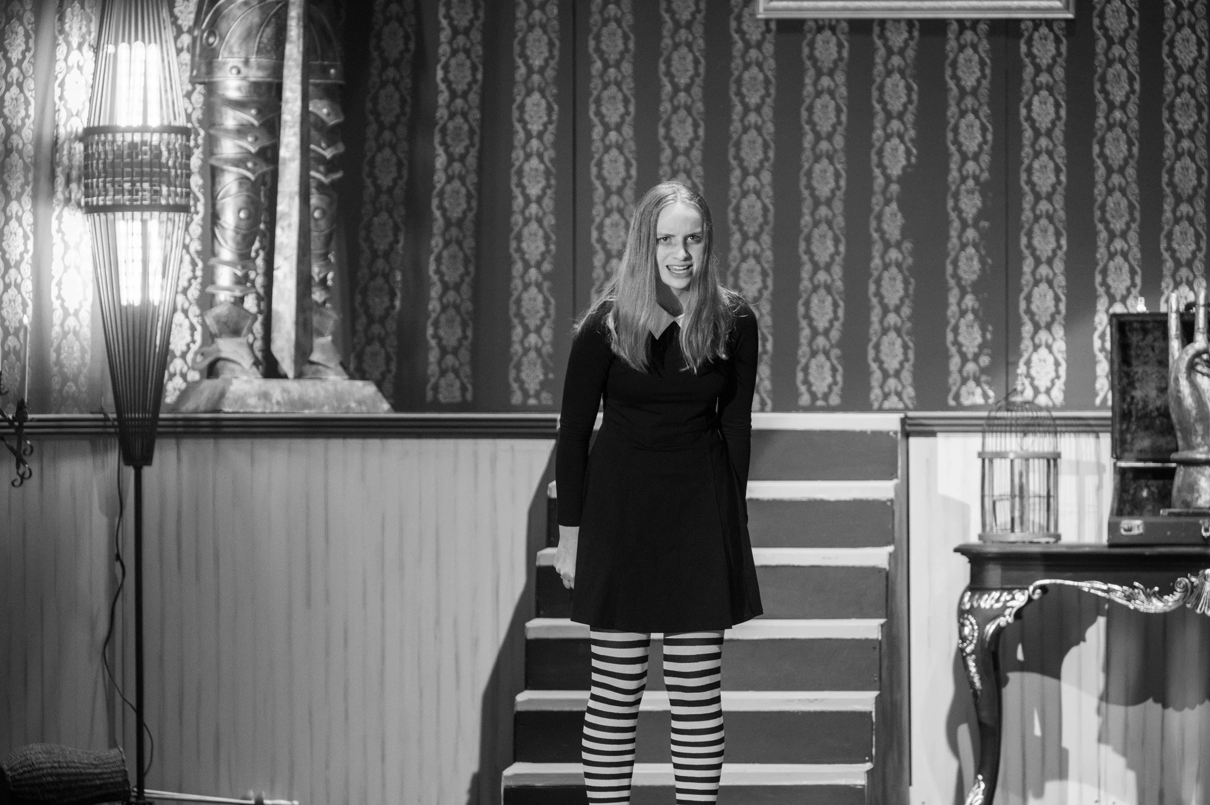 A woman with long hair wearing a dark dress and striped tights stands on a small staircase in a room with patterned wallpaper, a lamp, and vintage furniture.