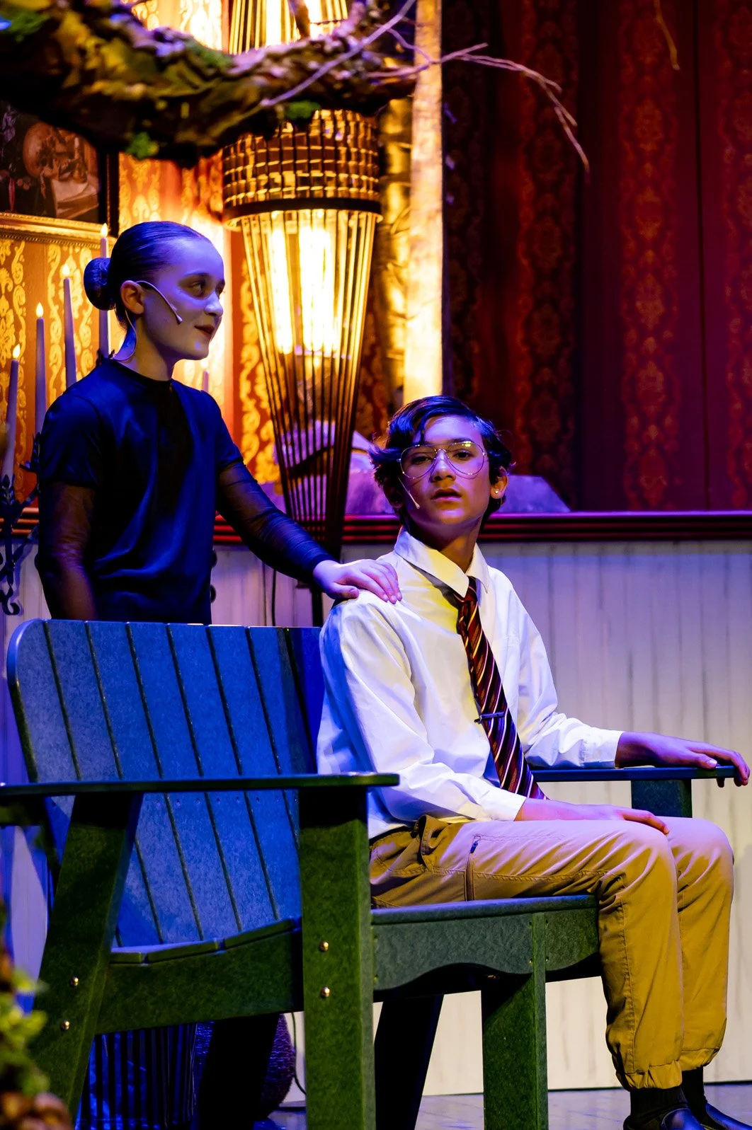 Two children on stage, a girl standing and a boy sitting on a bench, in a theatrical setting with warm and colorful lighting, ornate curtains, and vintage decor.