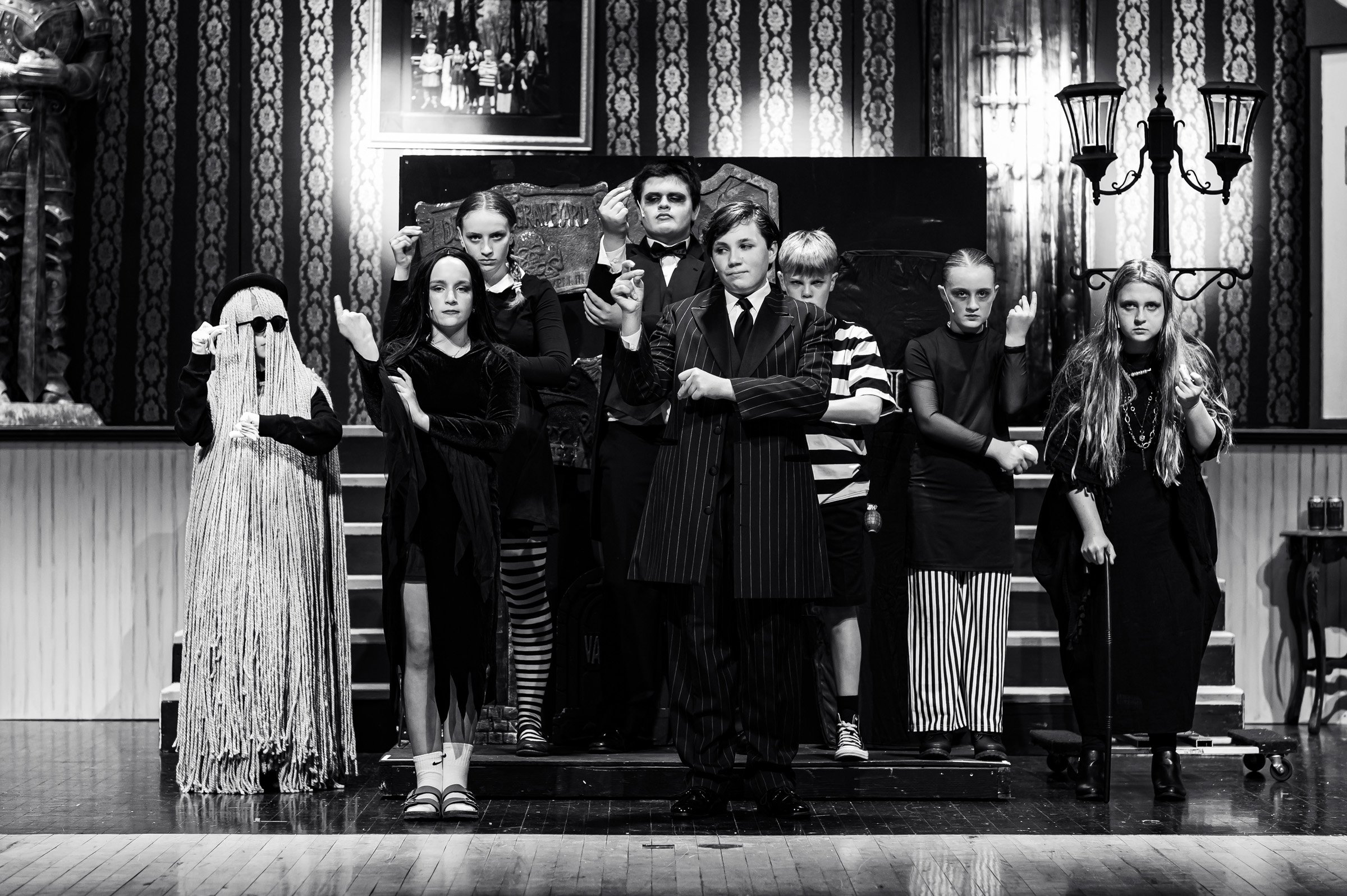 Black and white photo of children in Halloween costumes on a stage, with a patterned wallpaper background, a framed picture, and a street lamp. The children are dressed as various spooky characters, some with serious expressions.