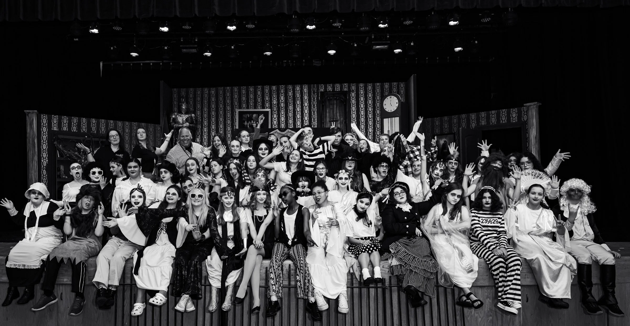 A large group of children and adults dressed in costumes for a theatrical performance, posing for a group photo on stage with a decorated background.