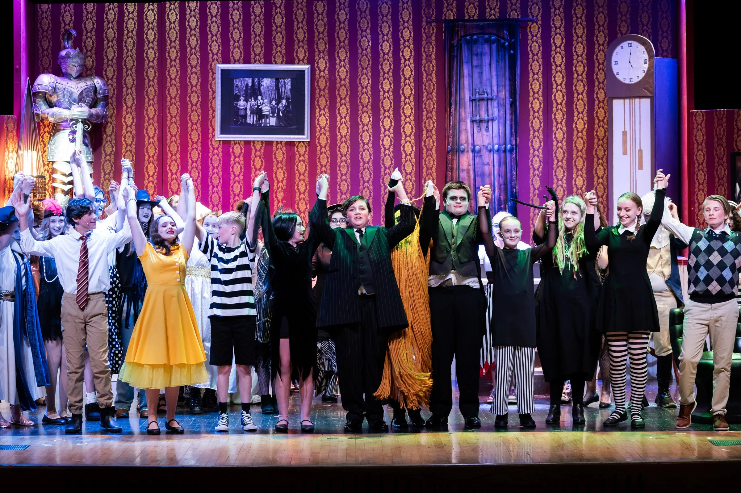 Group of children in costume on stage, holding hands and taking a bow after a theatrical performance, with a red ornate backdrop and theatrical props.