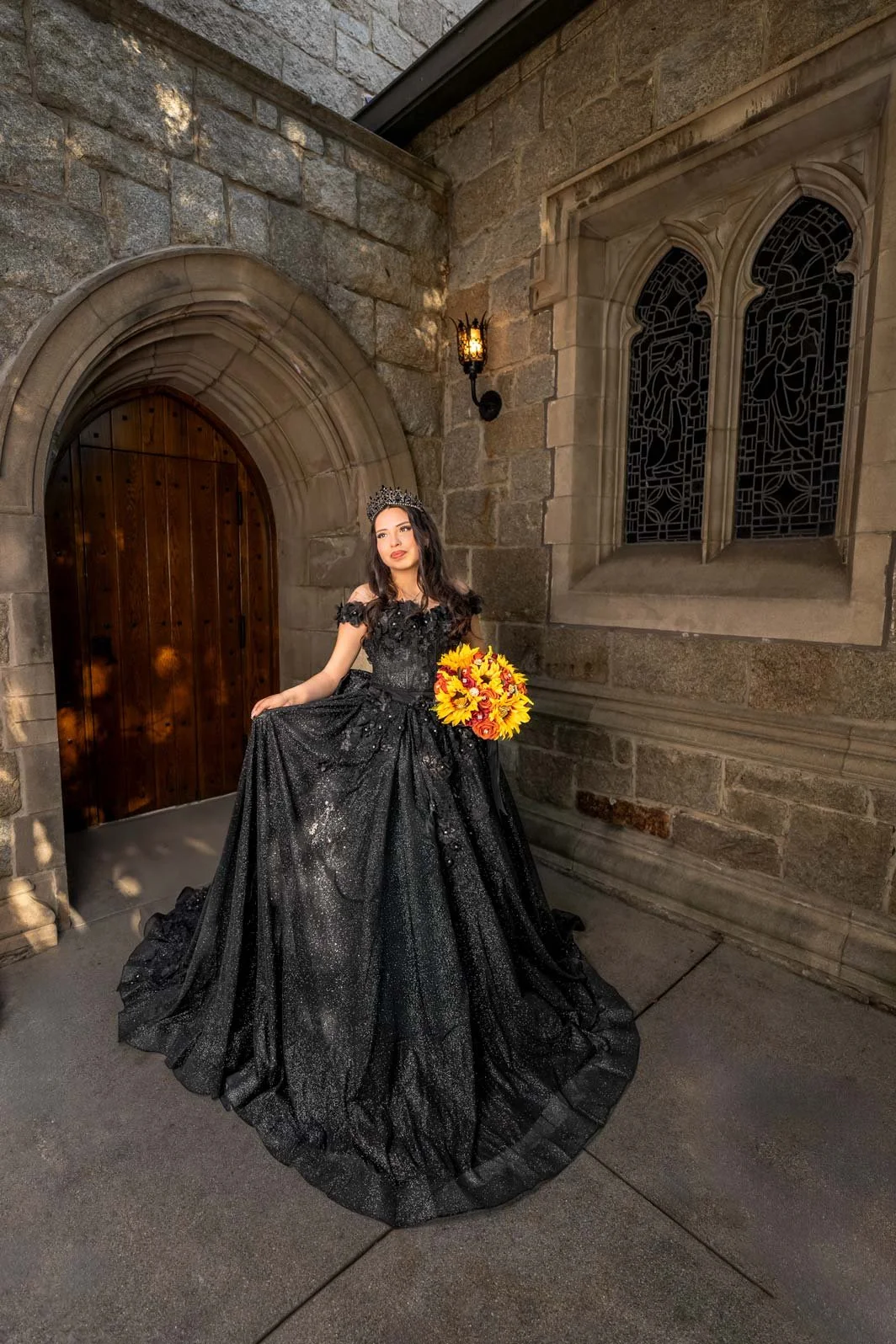 A woman in a black evening gown and tiara holding a bouquet of yellow and orange flowers stands outside a stone building with arched wooden door and stained glass windows.