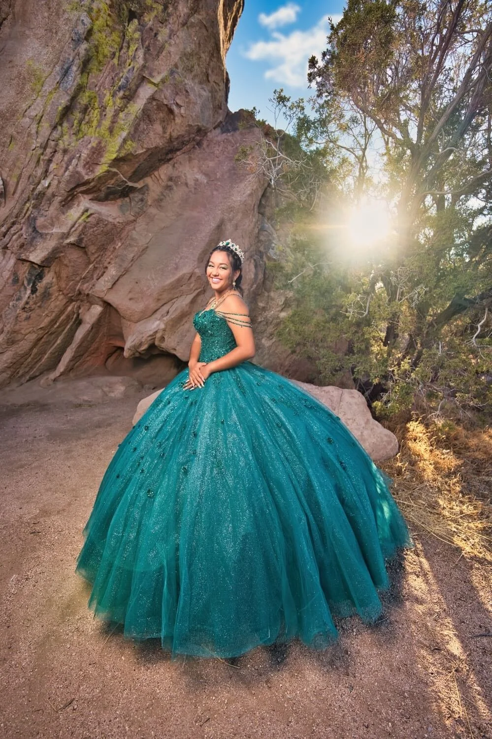 A young woman in an emerald green, sparkly ball gown and a tiara stands outdoors near rocks and trees with the sun shining in the background, smiling at the camera.