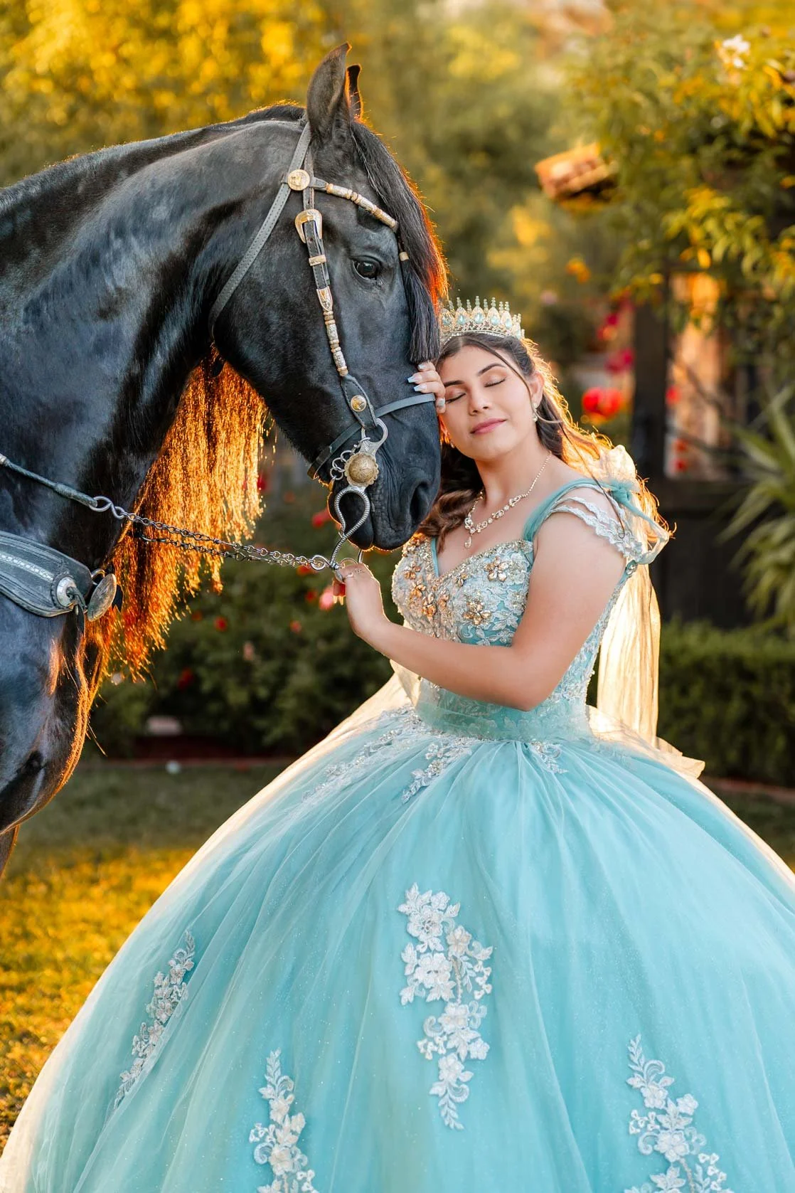 A young woman in a light blue ball gown with floral embroidery, wearing a tiara and necklace, gently cuddling a black horse in a garden during sunset.