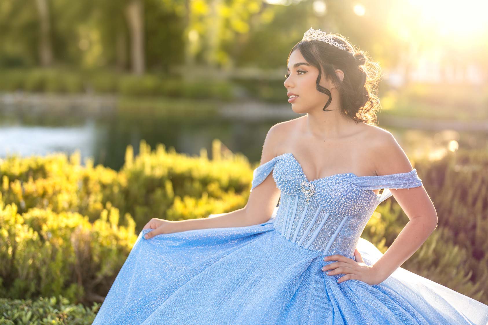A woman in a blue, sparkly off-the-shoulder gown with a tiara, standing outdoors near water with sunlight in the background.