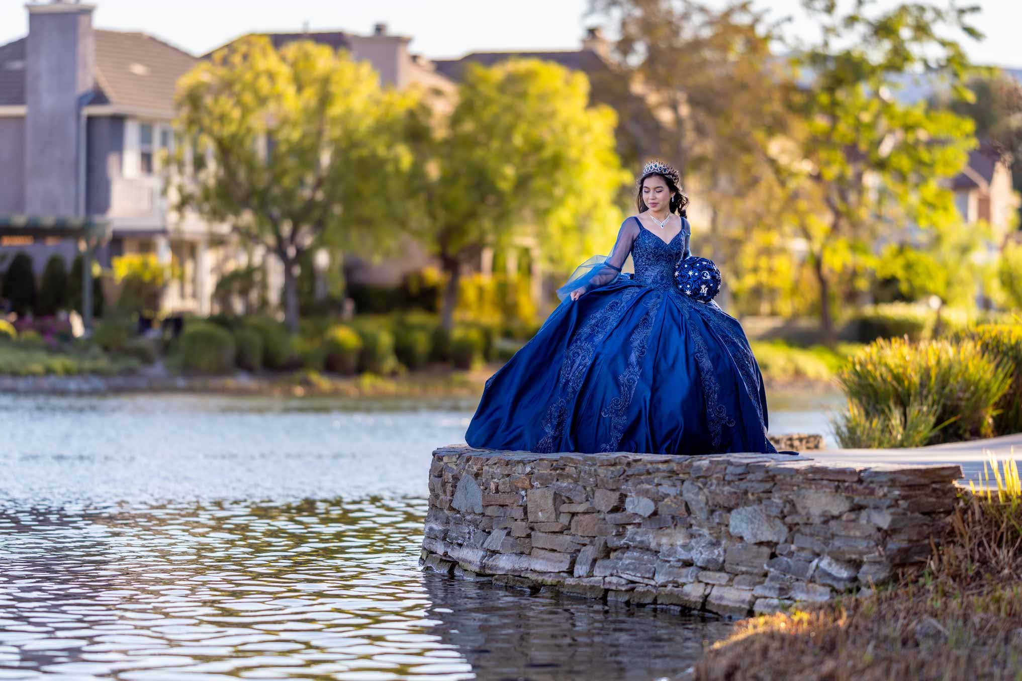 A woman in a blue ball gown with a tiara and jewelry standing on a stone ledge by a pond, with trees and houses in the background.