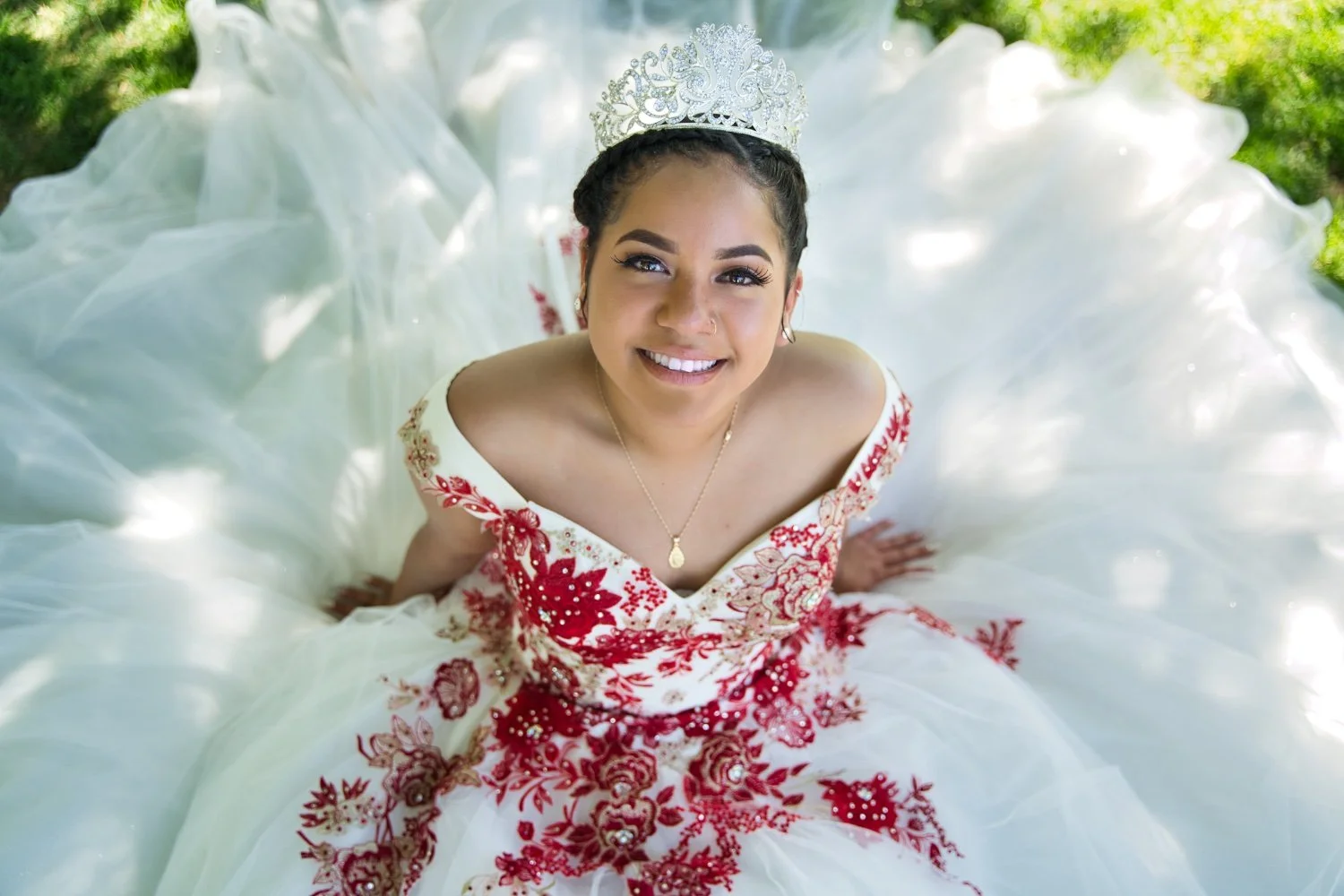 Young woman in a white and red embroidered dress with a tiara, smiling and looking up at the camera, sitting outdoors on the grass with a flowing, layered skirt spread around her.