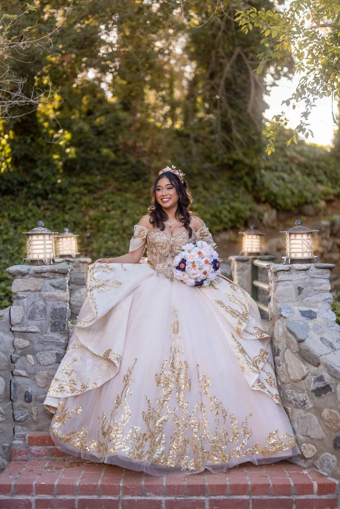 A bride in a gold and white wedding gown holding a bouquet of white, purple, and pink flowers, standing outdoors on a red brick platform with a stone railing and lanterns, surrounded by trees and greenery, smiling.