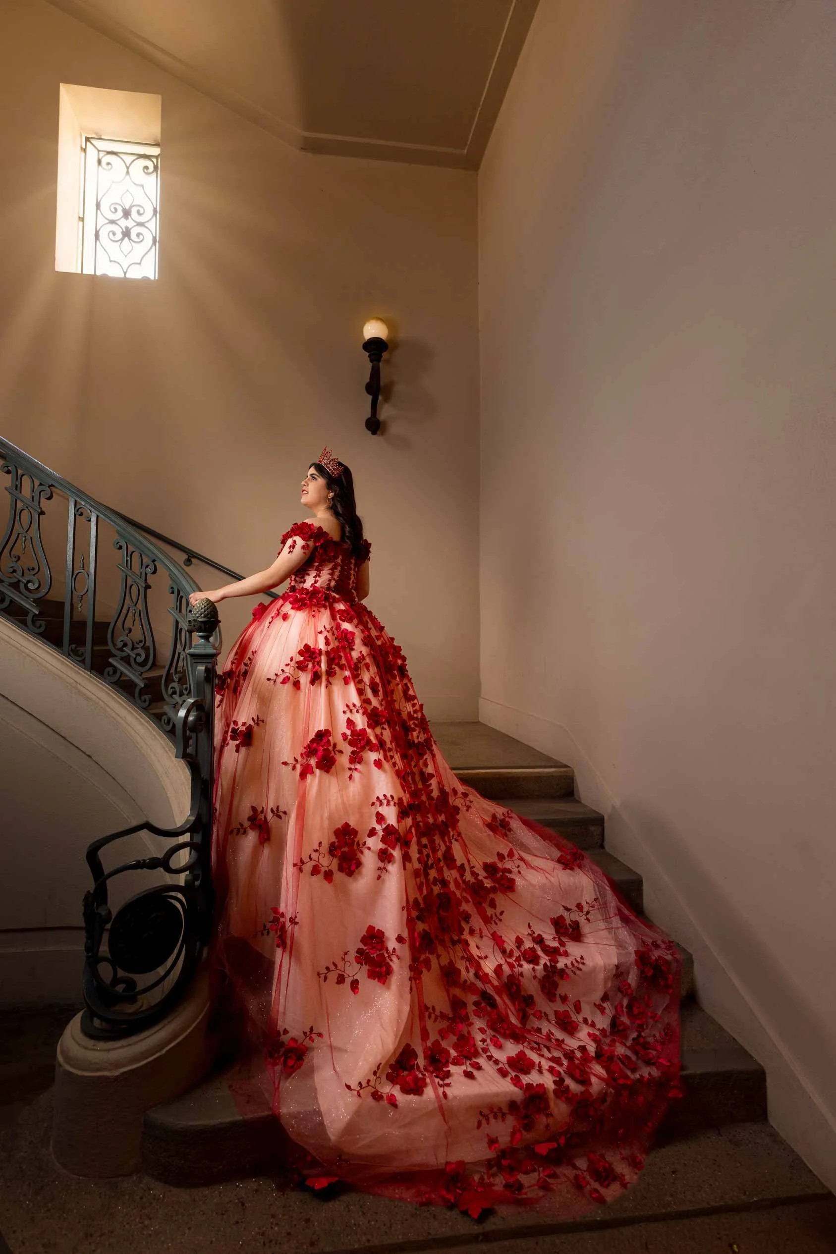 A woman in a red floral gown standing on a staircase in an interior setting with a wall light and a window casting shadows.