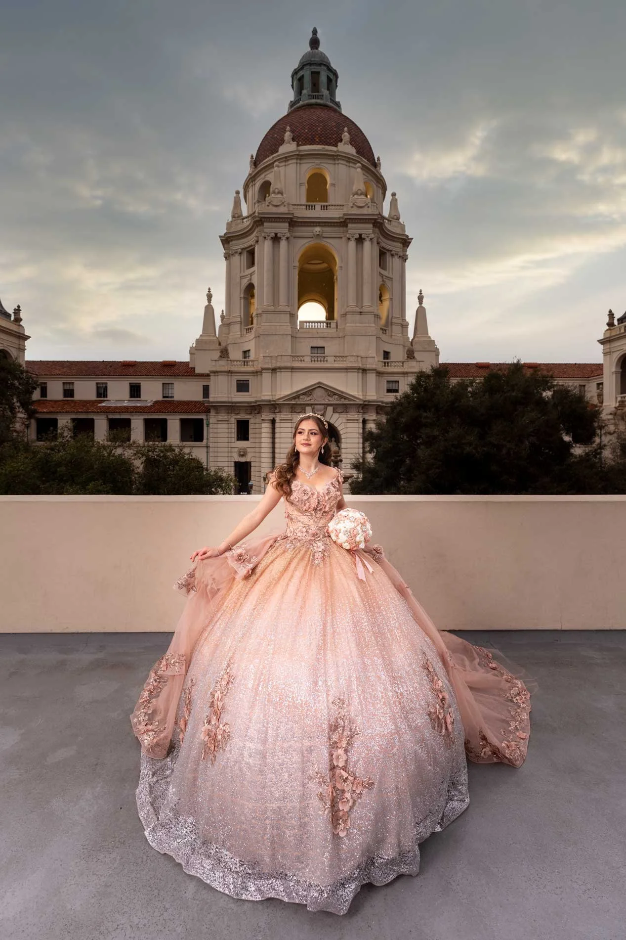 A young woman in a pink ball gown holding a bouquet stands on a rooftop with an ornate building with a large dome in the background, during sunset.