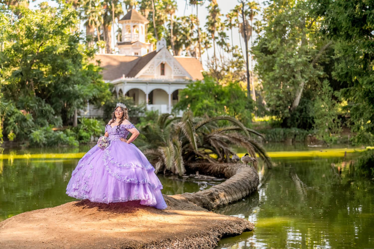 A young woman dressed in a lavender ball gown and wearing a tiara stands on a large rock by a pond, holding a bouquet, with a castle-like house and tall trees in the background.