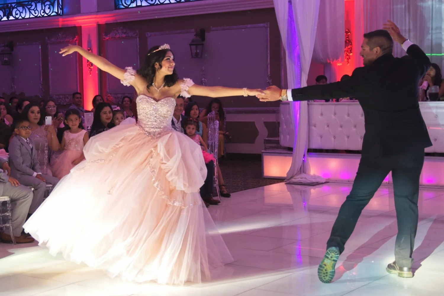 A bride and groom dancing at their wedding reception, with guests seated in the background watching and taking photos.