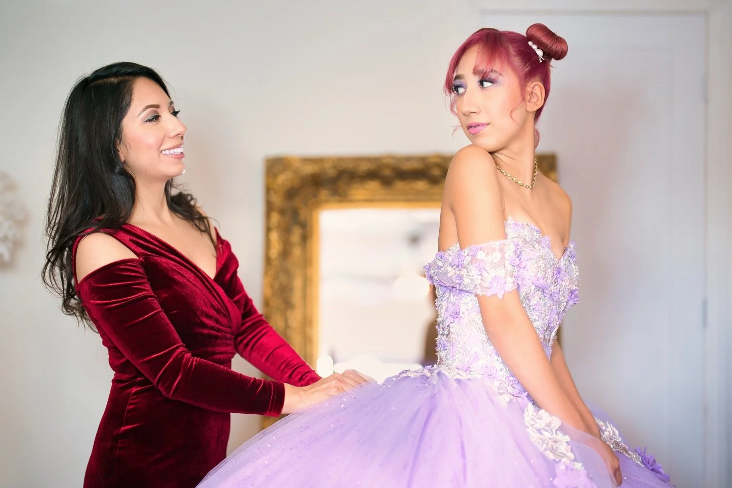 Two women, one in a red velvet dress and one in a lavender ball gown, smiling at each other indoors, with a mirror and picture frame in the background.