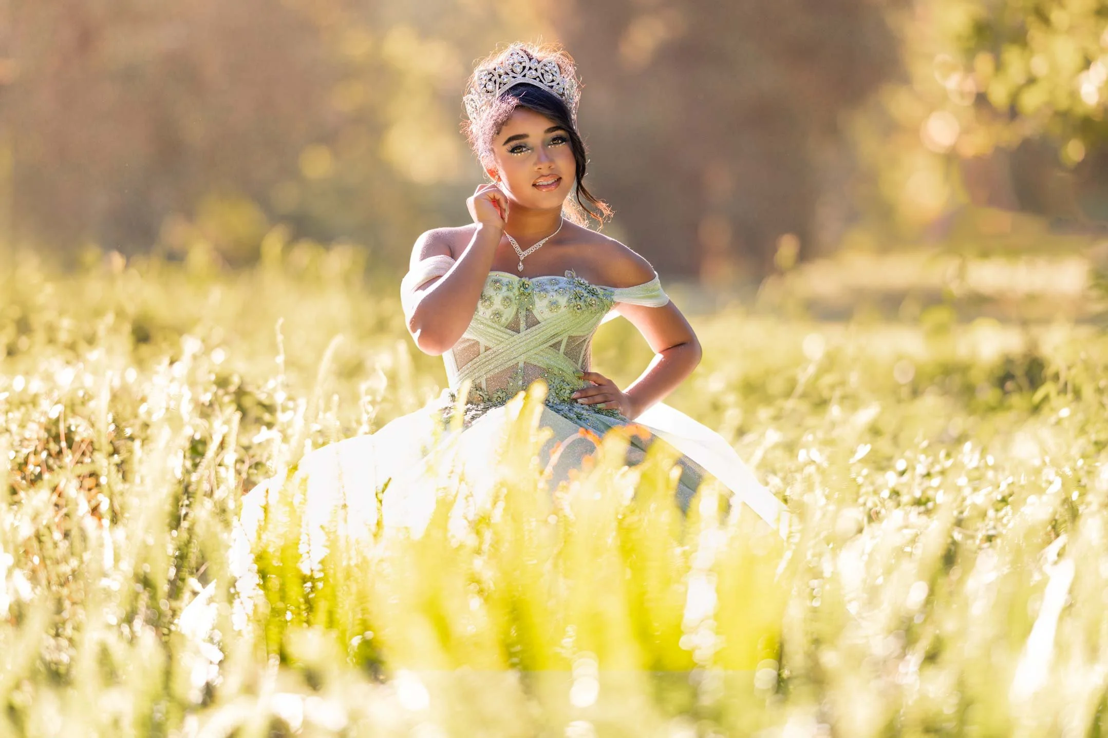A woman in a off-shoulder, silver and pastel green ball gown and a tiara, standing in a yellow flower field during golden hour, holding her chin with one hand, and looking at the camera.