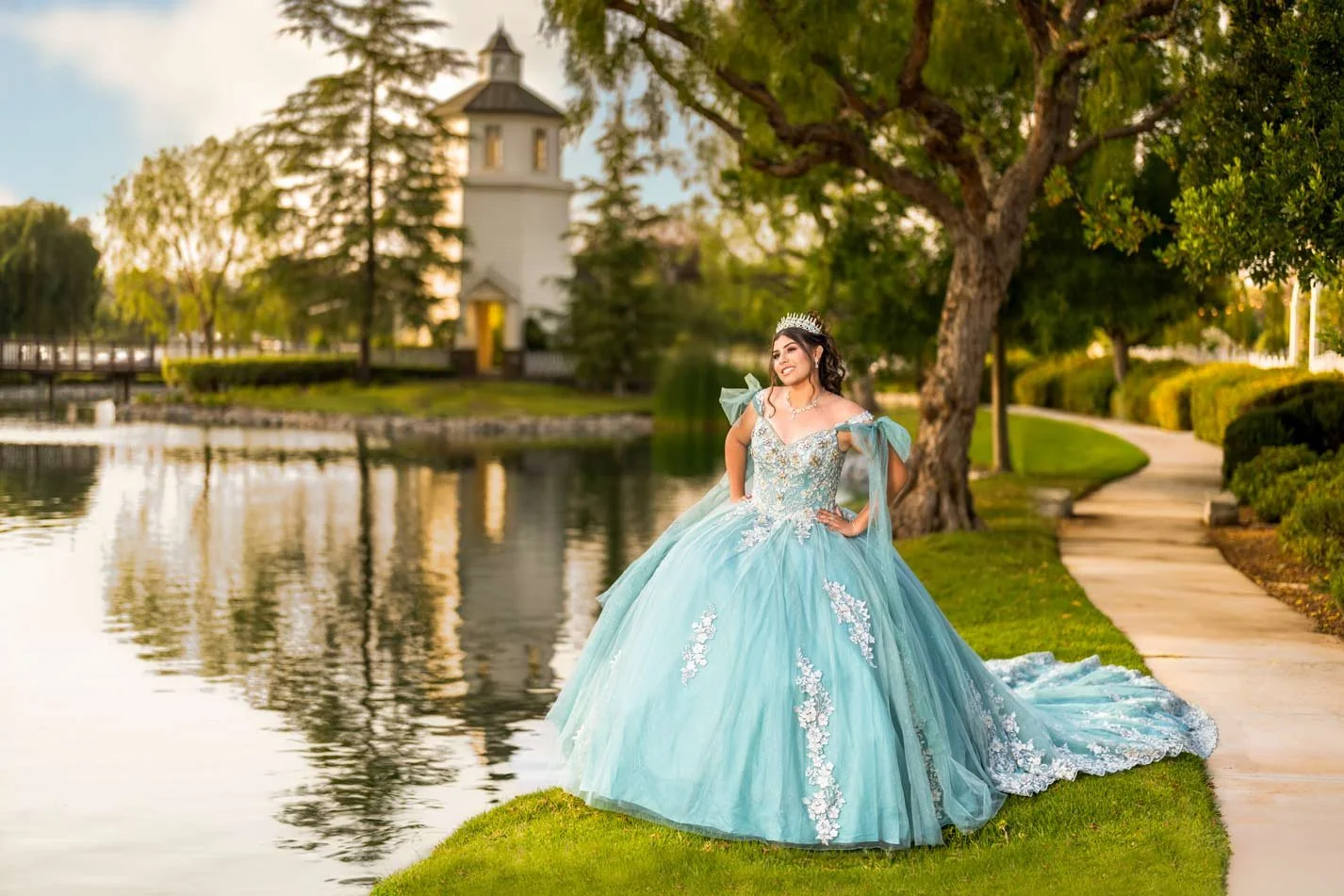 Young woman in a light blue ball gown with floral embroidery, wearing a tiara, standing by a lake on a grassy area with trees and a pathway, during sunset.