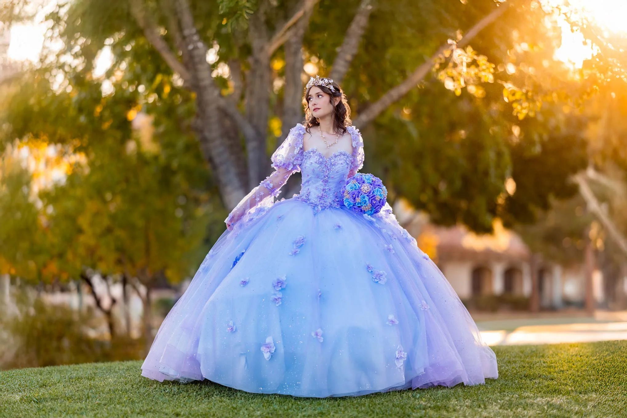 Young woman in a light blue ball gown holding a bouquet of blue roses, standing outdoors near trees in the golden hour sunlight.