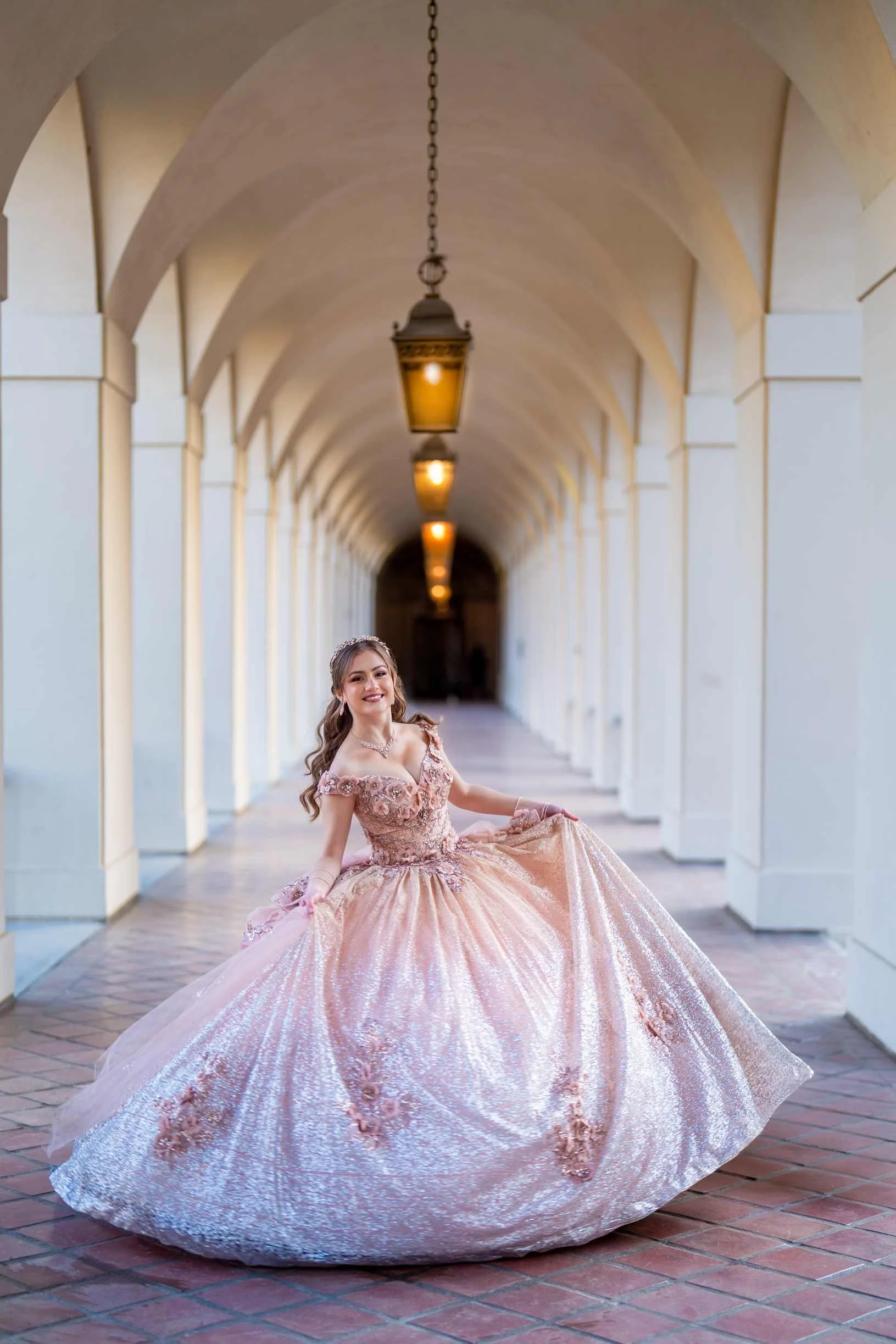 A young woman in a pink ball gown with floral designs, standing in an arched hallway with hanging lanterns, smiling and holding out her skirt.