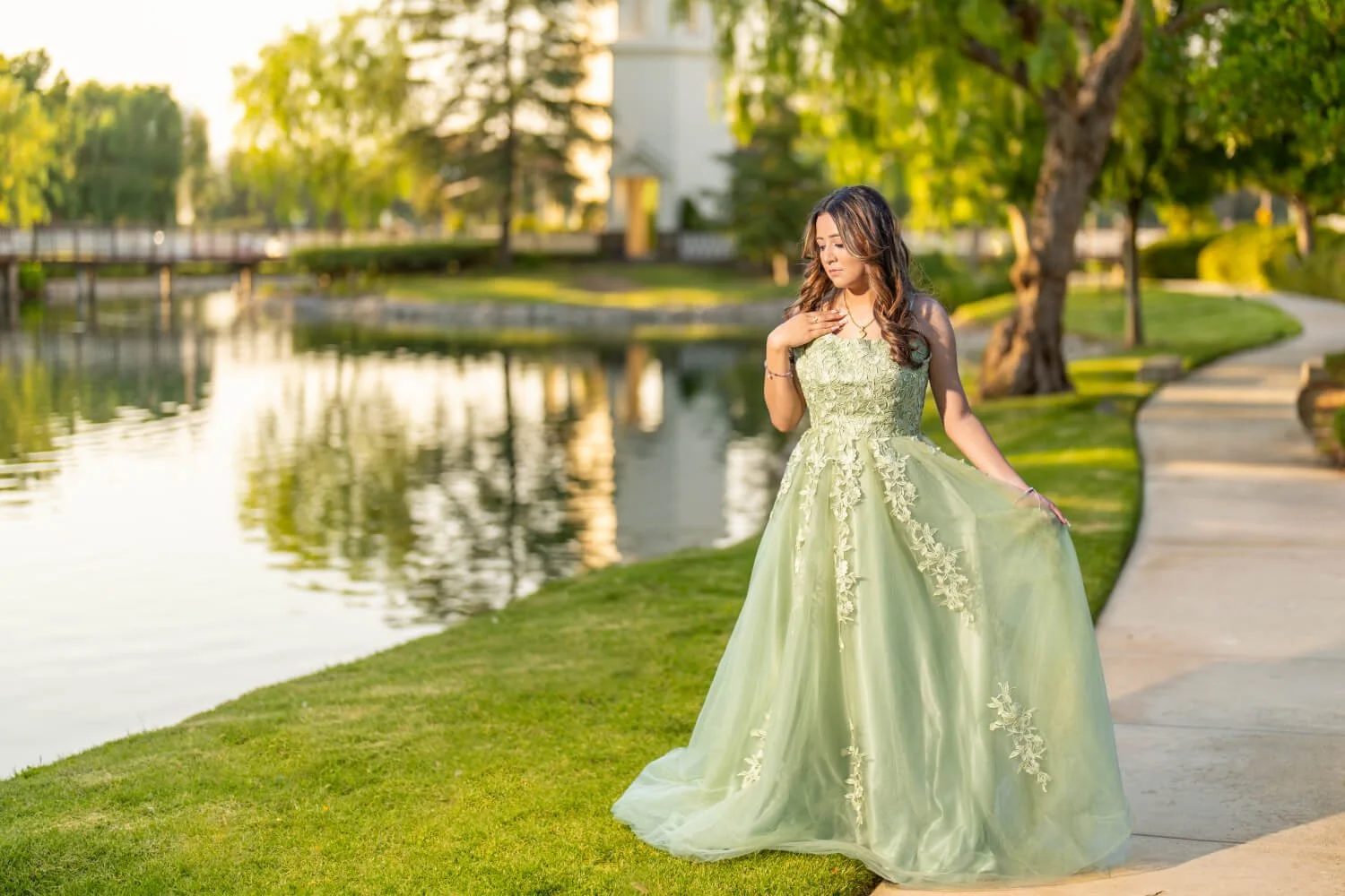 A young woman in a green embroidered gown standing by a river in a park during sunset, with trees and a walking path in the background.