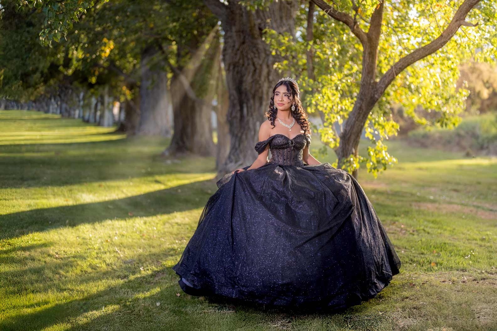 Young woman in a black, sparkly ball gown standing outdoors in a park with trees and grass, during golden hour sunlight.