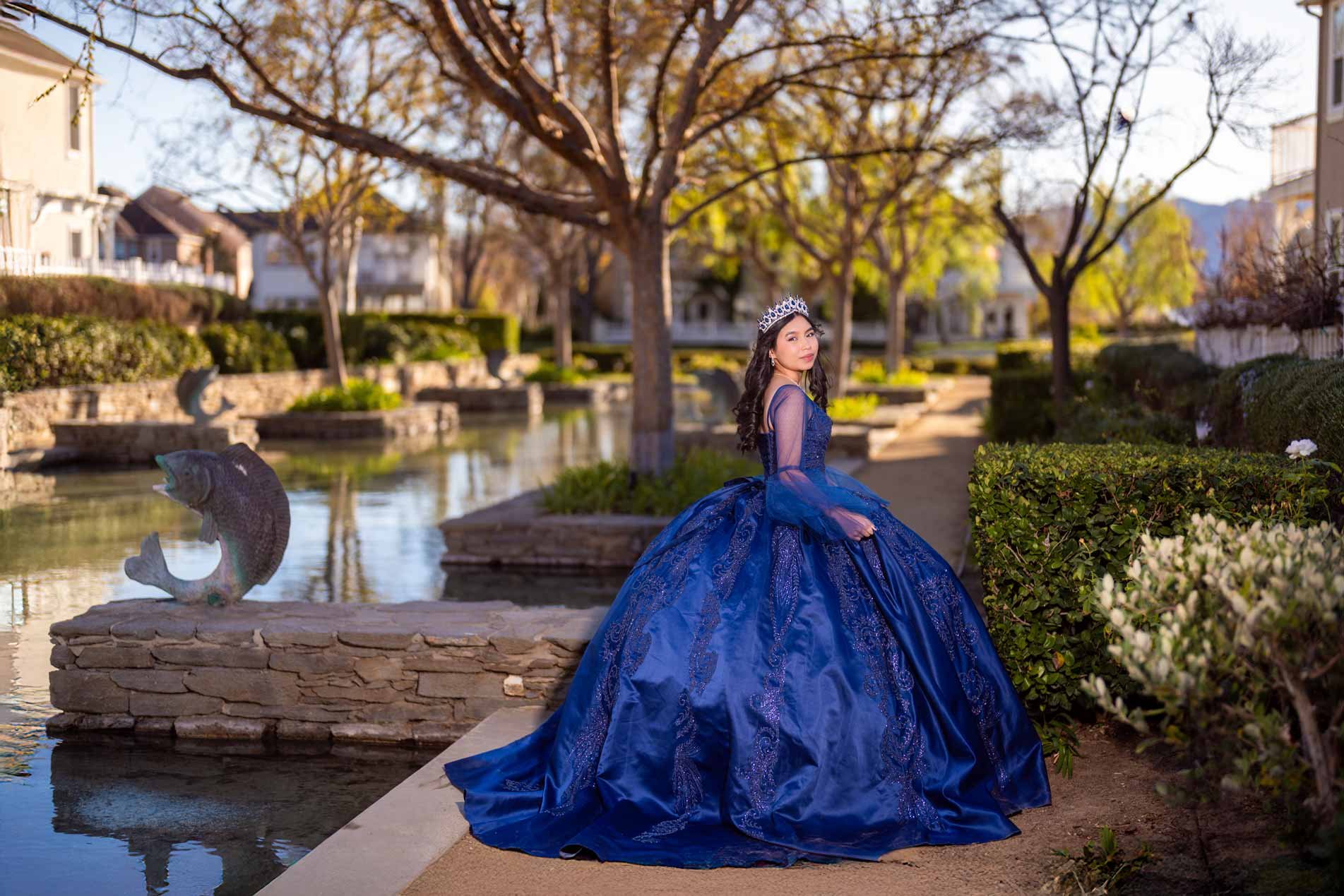 A young woman in a royal blue ball gown and tiara standing by a pond with fish sculptures, trees with autumn leaves, and residential houses in the background.