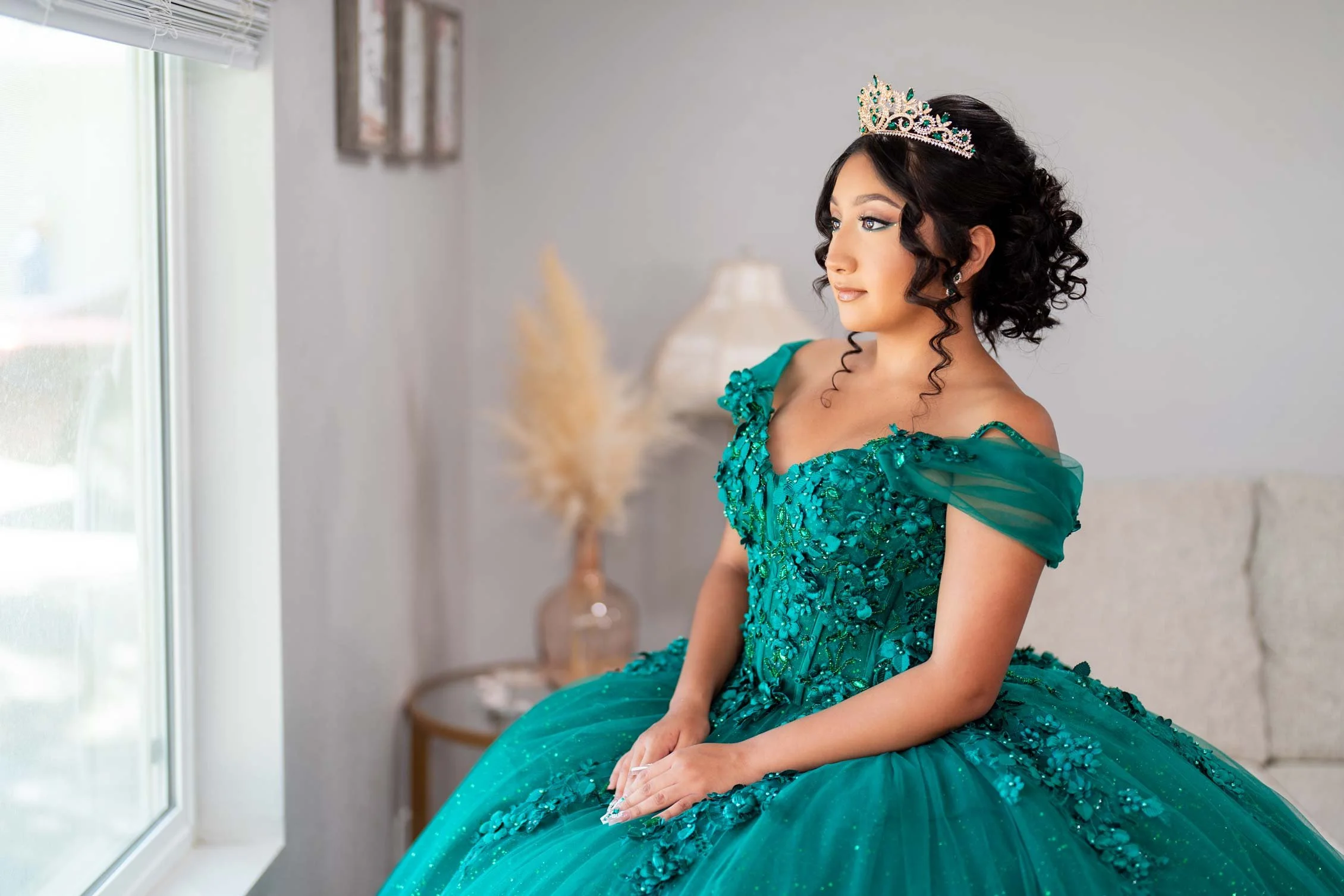 Young woman in an elaborate teal gown with floral embellishments, sitting near a window with her eyes closed, wearing a tiara, with a blurred background of a lamp and a vase of pampas grass.