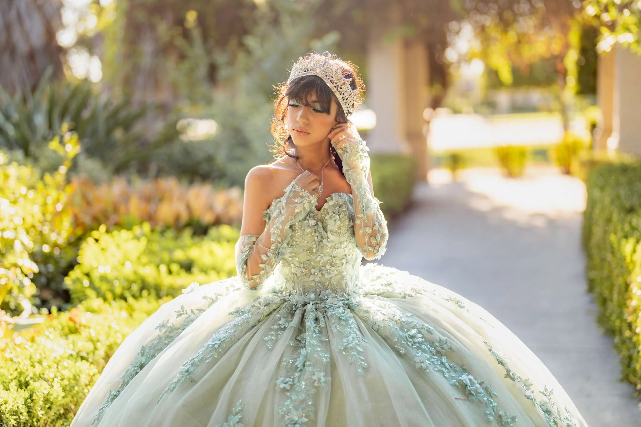 A young woman in an elegant, light green ball gown with floral embellishments and long lace gloves, standing outdoors in a sunlit garden, wearing a tiara, with her eyes closed and her hand gently touching her neck.
