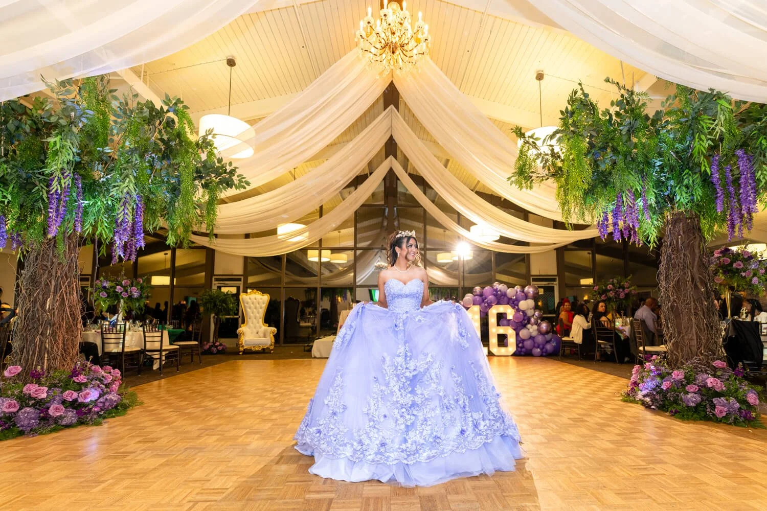 Bride in a lavender wedding gown standing on dance floor decorated with floral arrangements and purple balloons, under draped fabric and a chandelier in a reception hall.