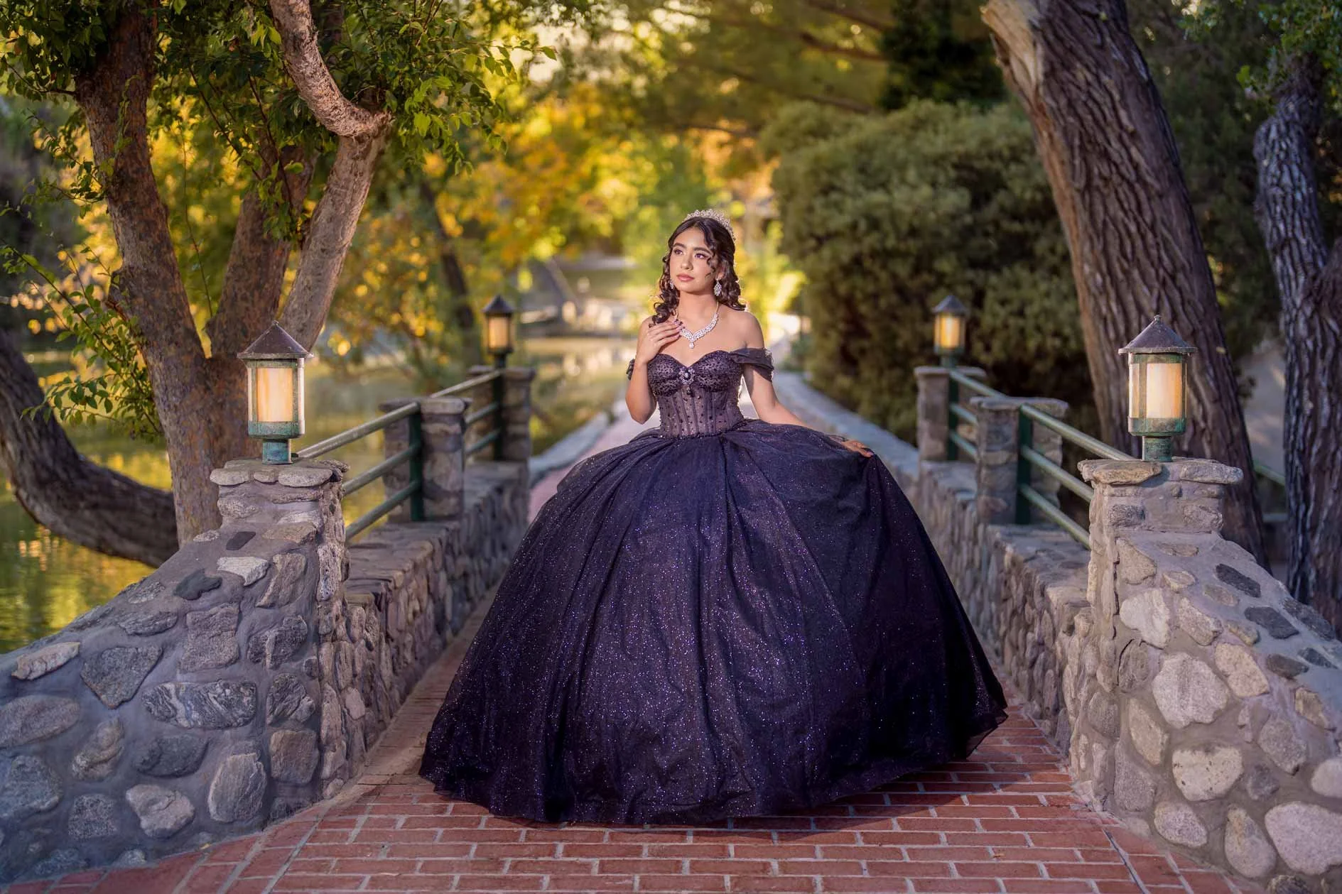 A woman in an elegant, sparkly, dark purple ball gown posing on a stone walkway with a railing, surrounded by trees and lanterns, during sunset.