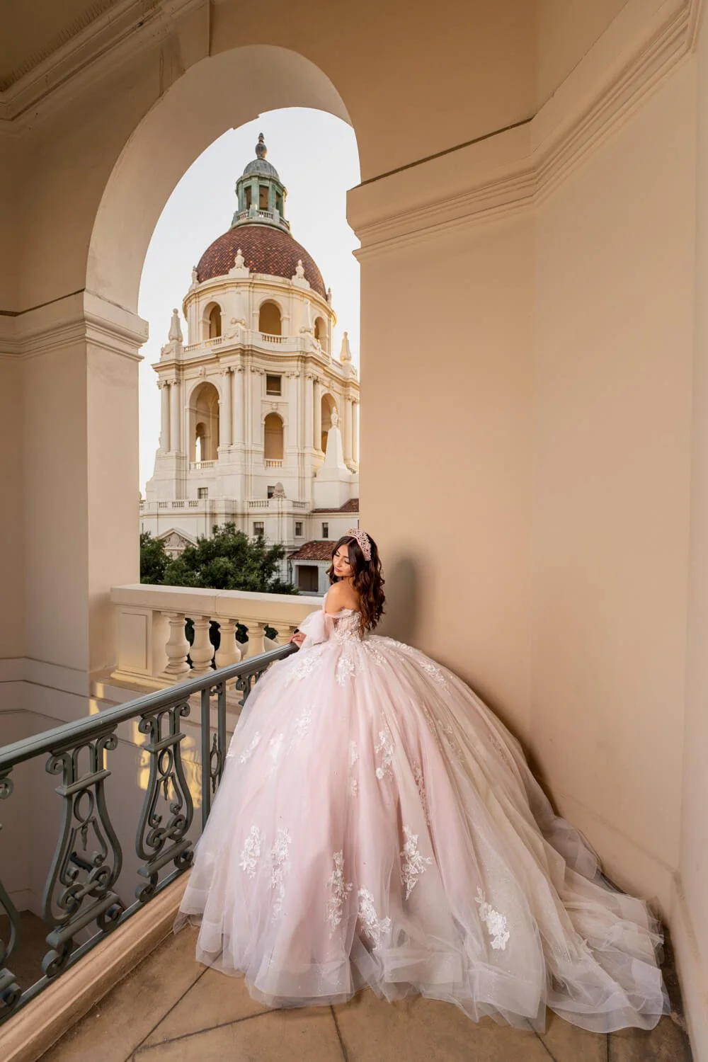 A woman in a pink lace wedding gown with a tiara on her head standing on a balcony with a view of a grand cathedral with a large dome in the background.