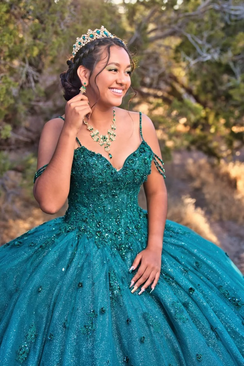 A young woman in a teal, beaded, and sequined ball gown with a full skirt, wearing a matching jewelry set and a tiara, outdoors with trees in the background.