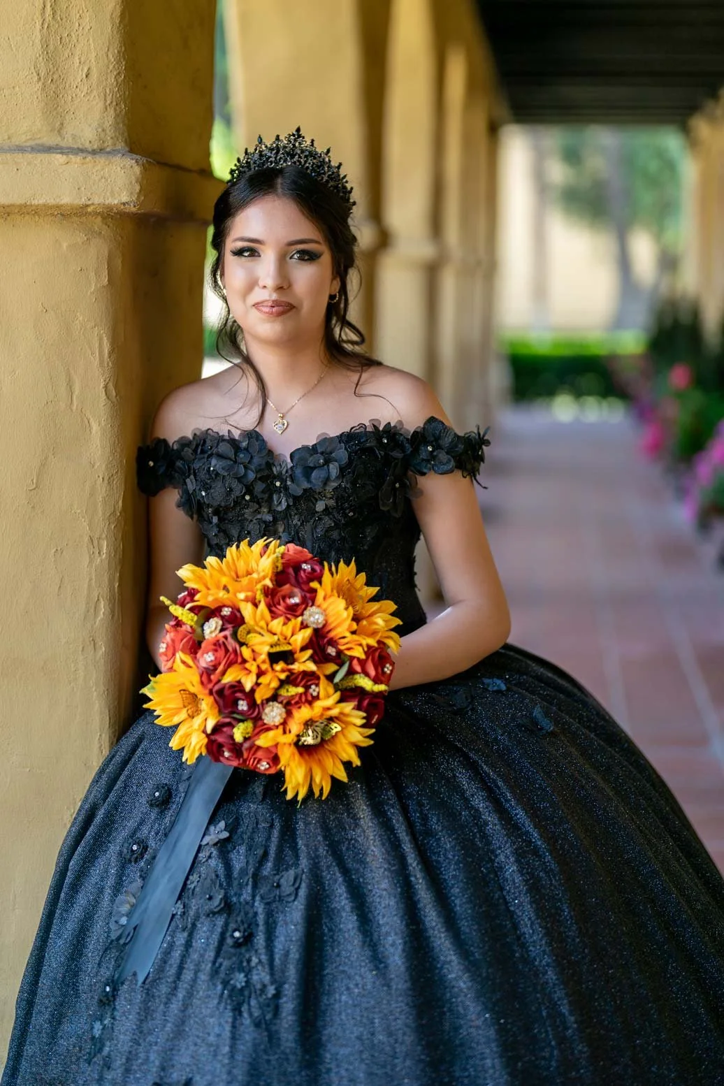 A young woman in a black off-shoulder gown with floral embellishments, holding a bouquet of sunflower and red roses, standing against a yellow stucco wall in an outdoor corridor.
