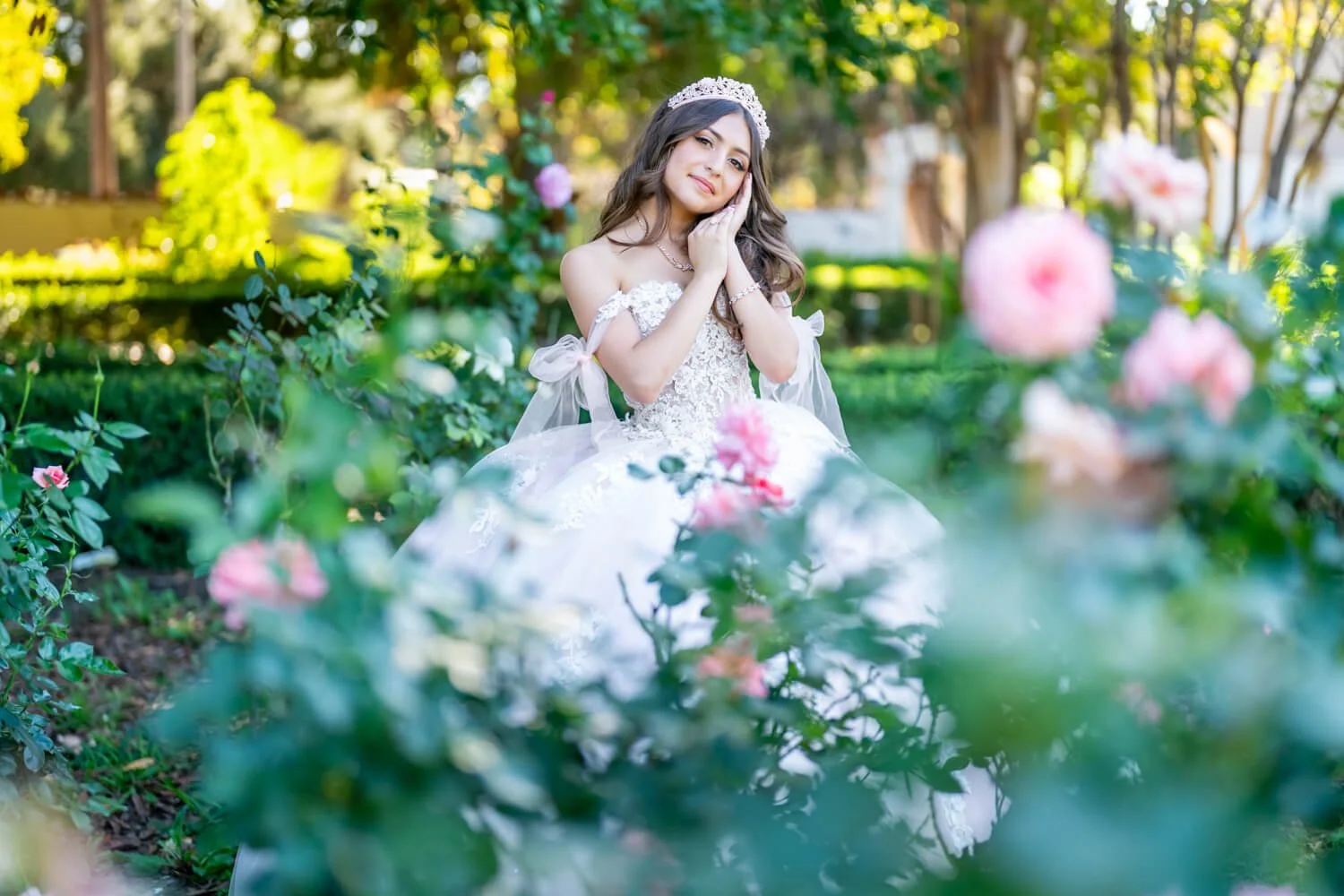 A young woman dressed in a white wedding gown with lace details, wearing a tiara, sitting among pink roses and greenery in a garden, with a gentle expression and her hands resting on her cheek.