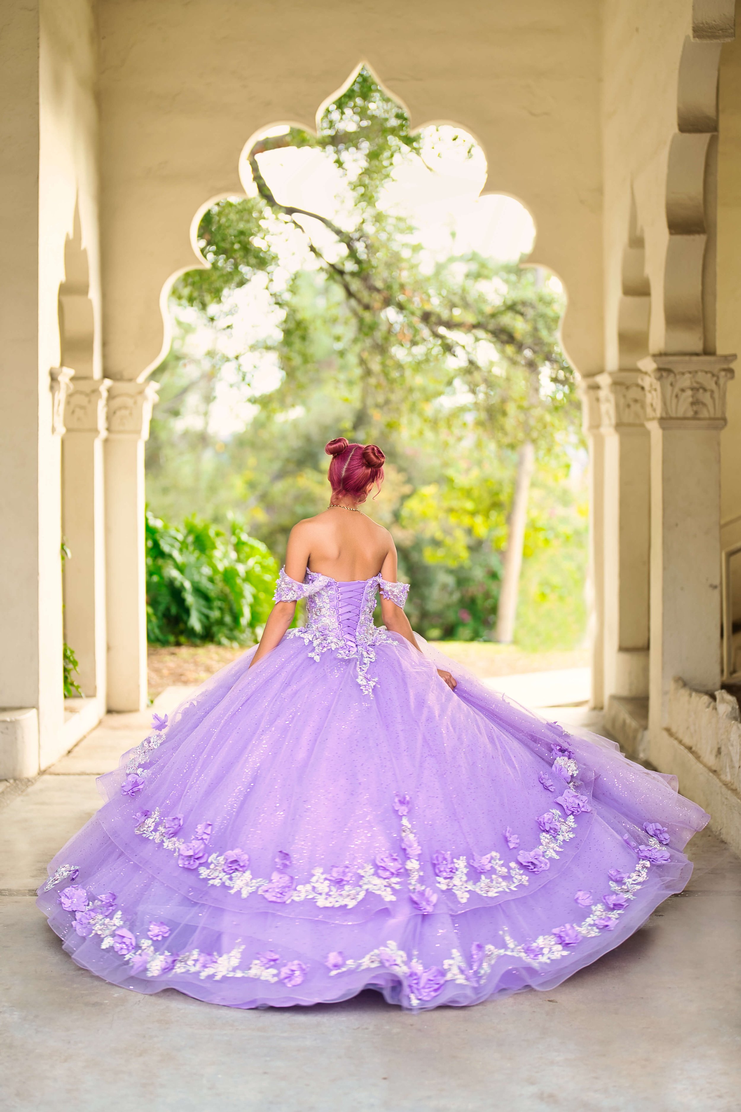 A woman in a lavender ball gown with floral embroidery standing in an arched stone doorway, facing away, holding the train of her dress, with lush greenery and trees in the background.