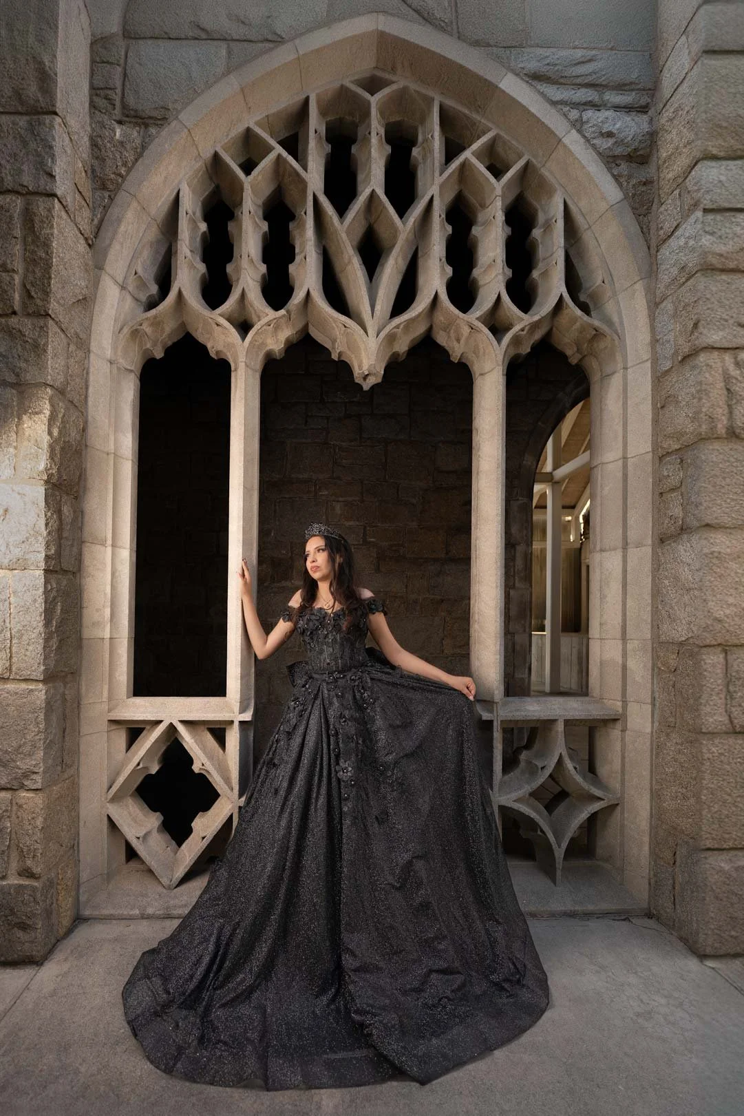 A woman in a long, black, off-the-shoulder gown with lace and embellishments, standing beneath a Gothic-style stone archway, holding part of her dress with one hand and touching the arch with the other.