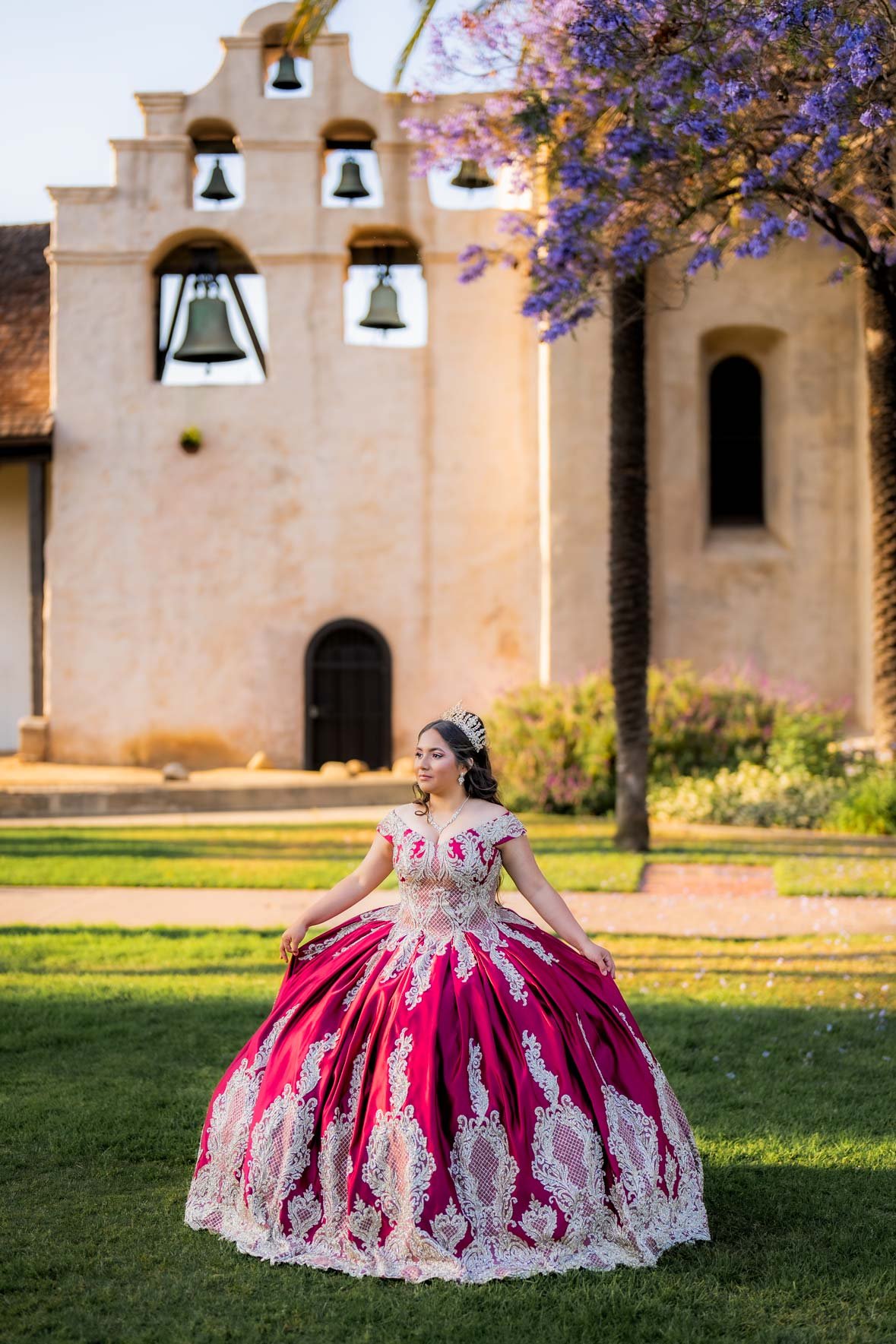 A woman in an elaborate pink and white gown standing on a lawn in front of a historic church with bells, with flowering trees in the background during daylight.
