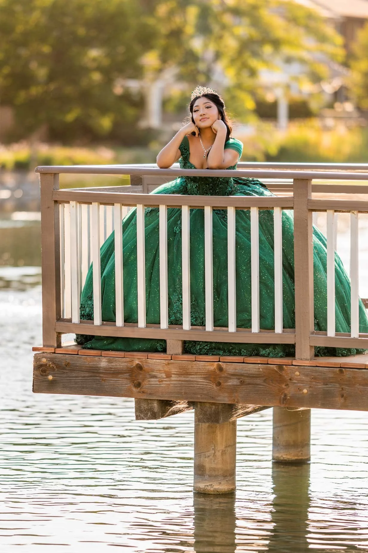 A young woman in a green prom gown with a tiara and necklace, standing on a wooden dock over water, with trees in the background.