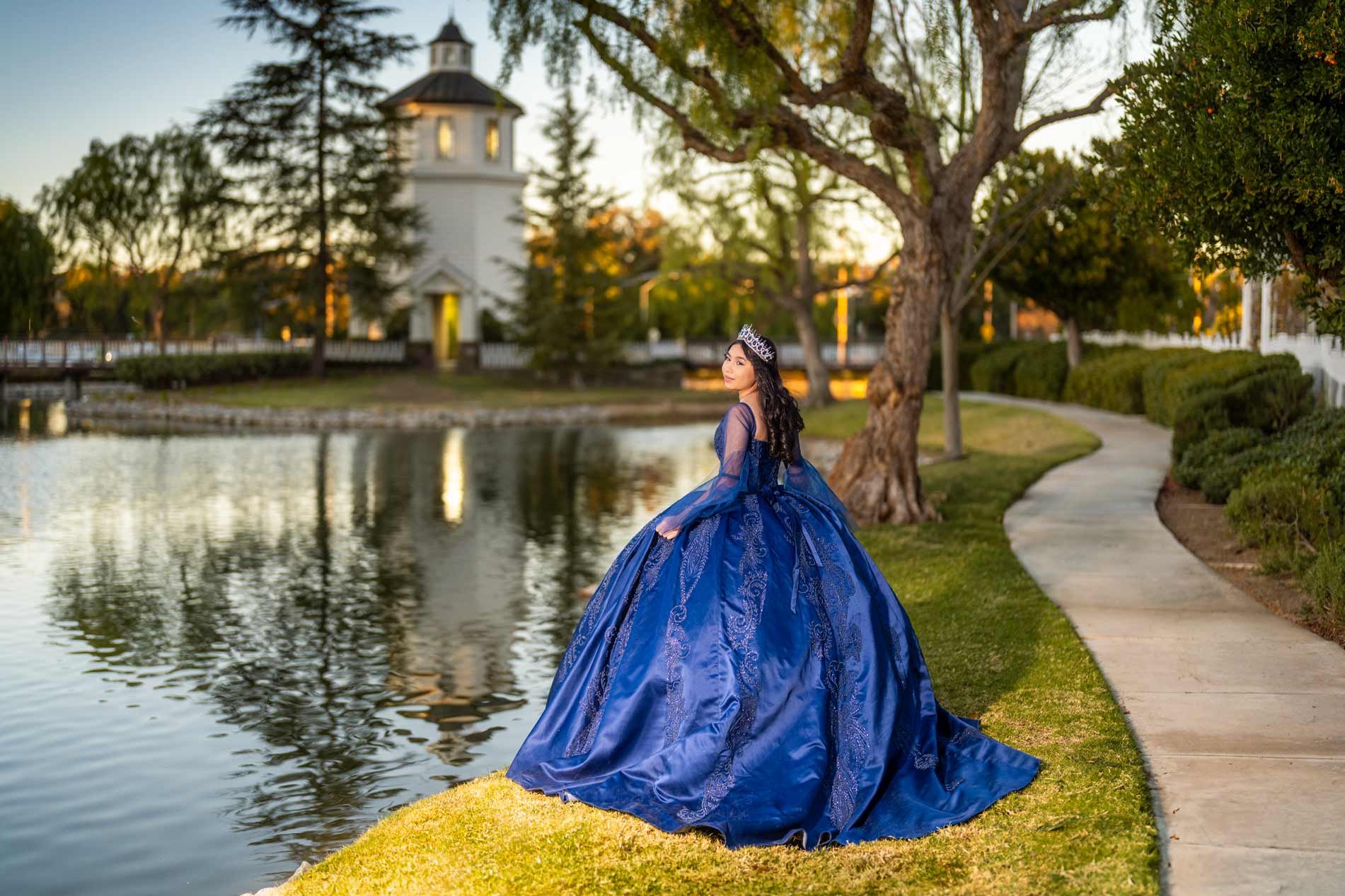 Young woman in a royal blue, ball gown with lace details, wearing a tiara, standing near a pond in a park during sunset.