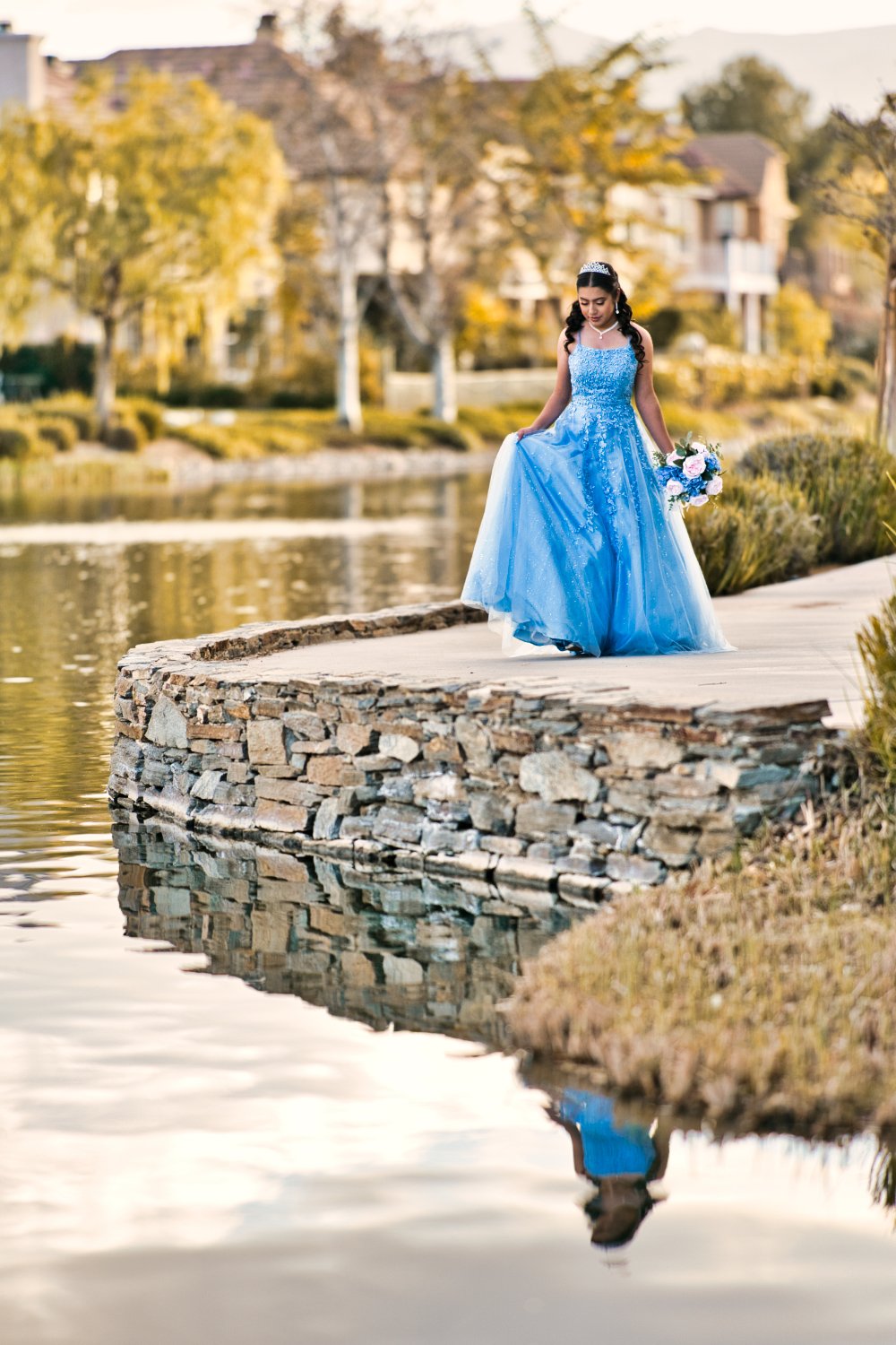 Young woman in a blue dress holding a bouquet standing on a stone dock by a river with trees and houses in the background.