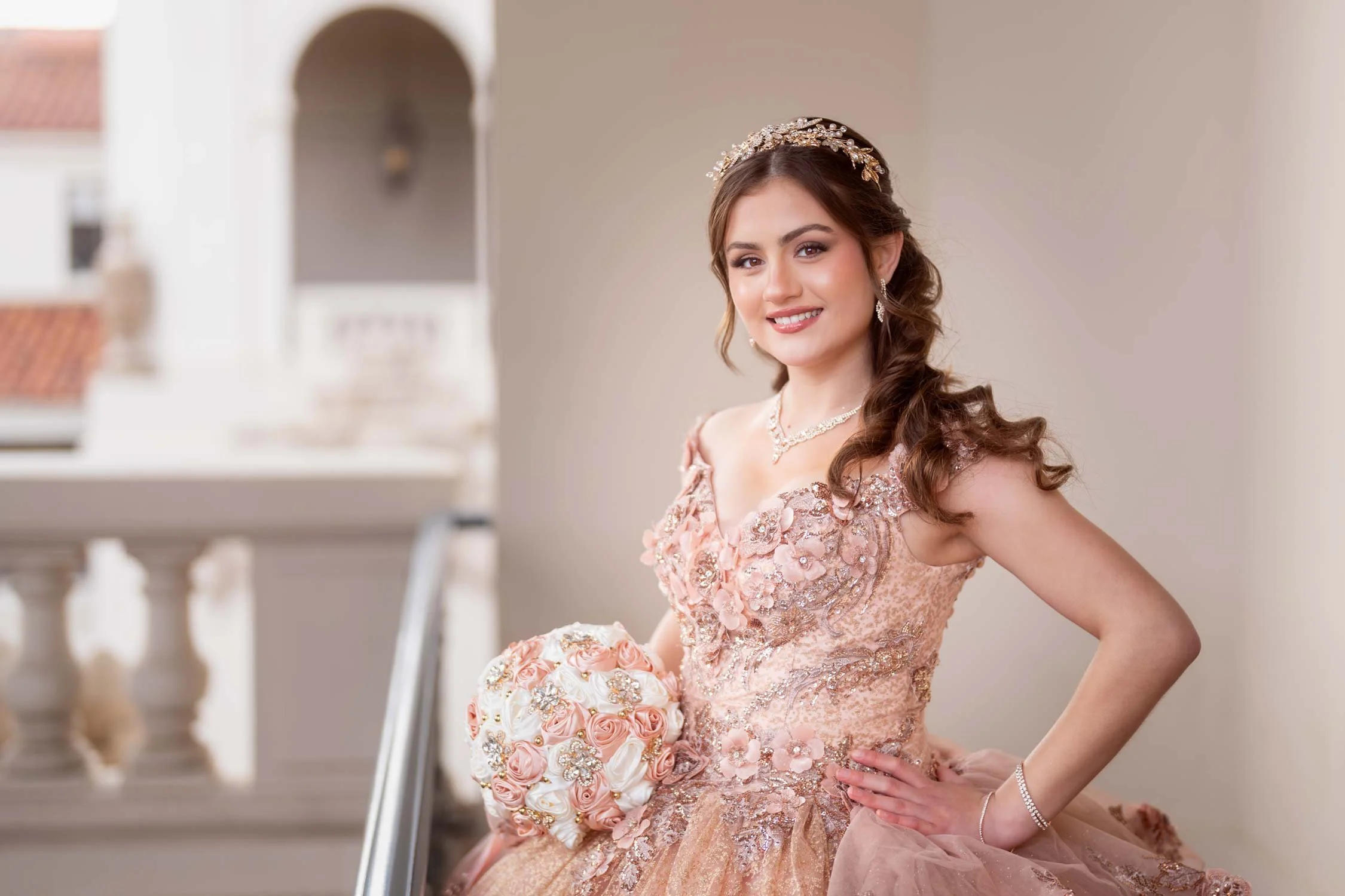 Young woman in an elegant peach lace dress holding a pink and ivory floral bouquet, standing on a balcony with a blurred background.