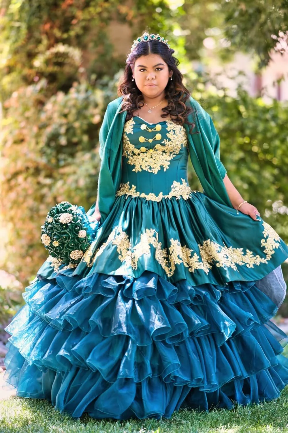 Young woman in a teal and gold formal dress, gold tiara, holding a teal bouquet, outdoors with greenery in the background.