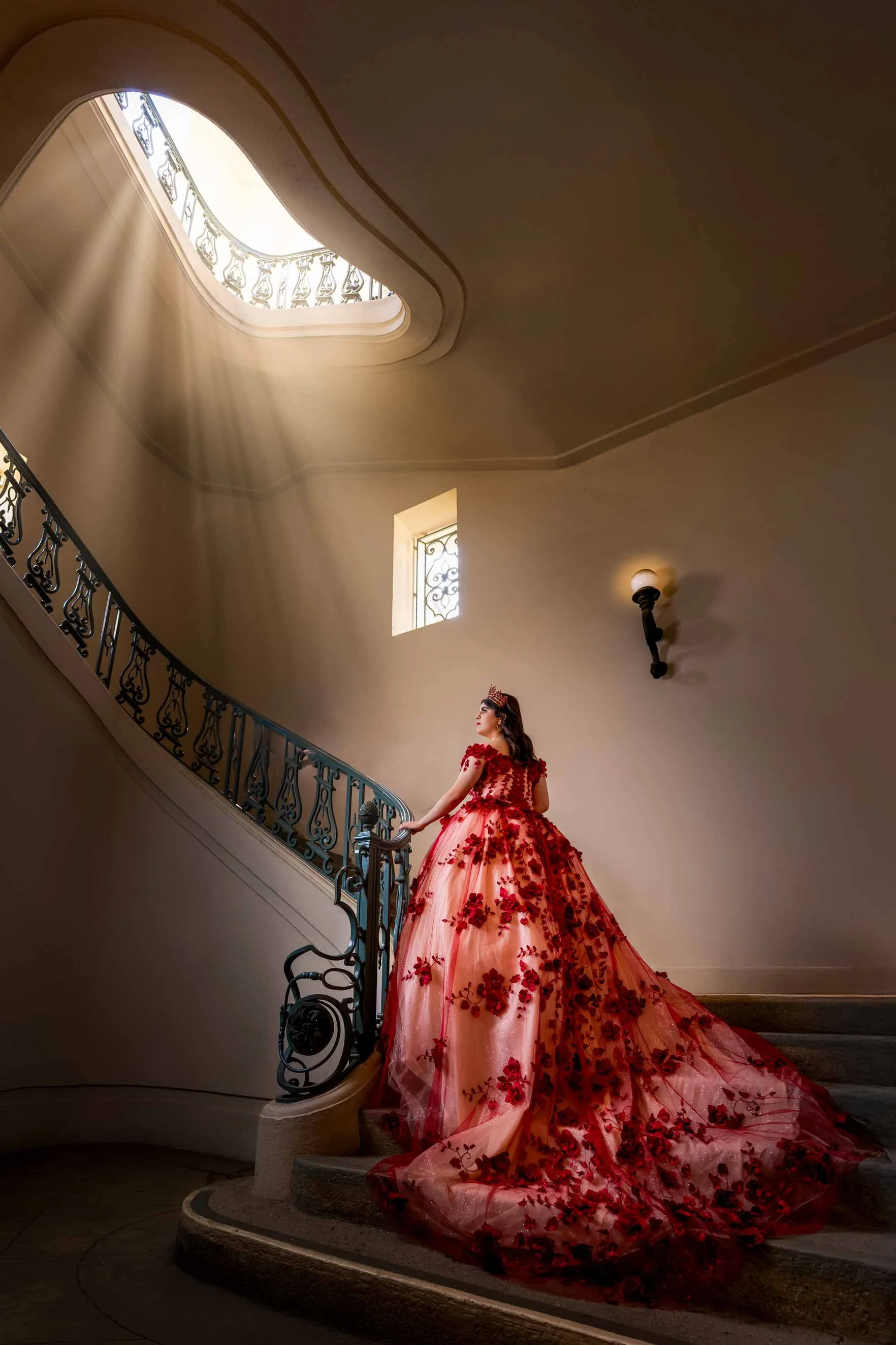 A woman in a red floral gown standing on a staircase with sunlight streaming through an oval skylight above.
