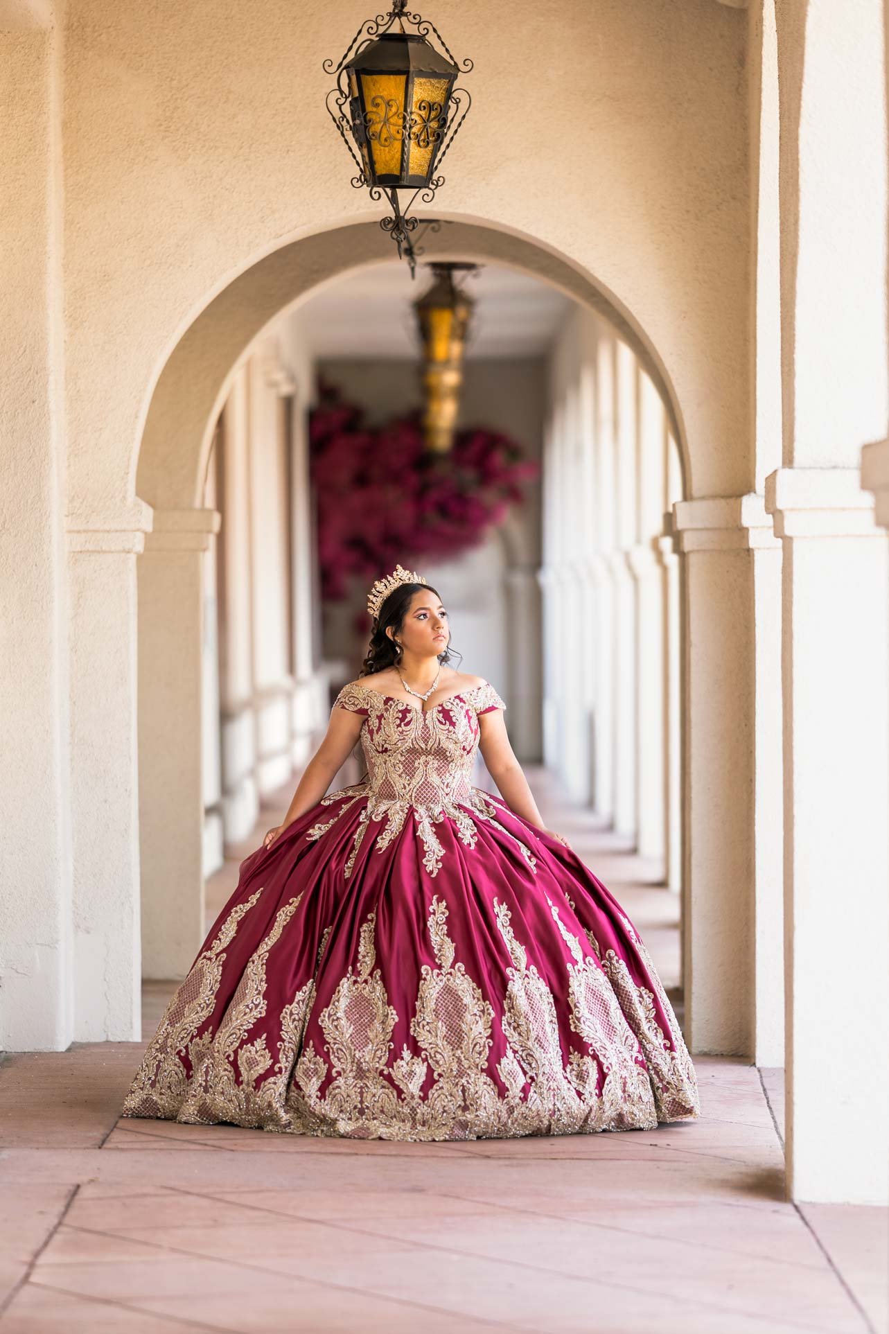 Young woman in an elaborate red and gold gown, wearing a tiara, standing in a hallway with arches and hanging lanterns.
