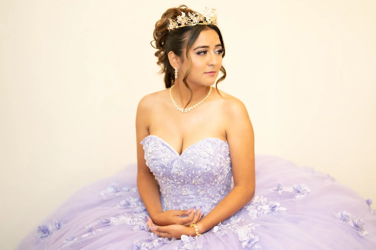 Young woman dressed as a bride, wearing a lavender wedding dress with floral embroidery, a tiara, pearl jewelry, and sitting against a plain light-colored wall.