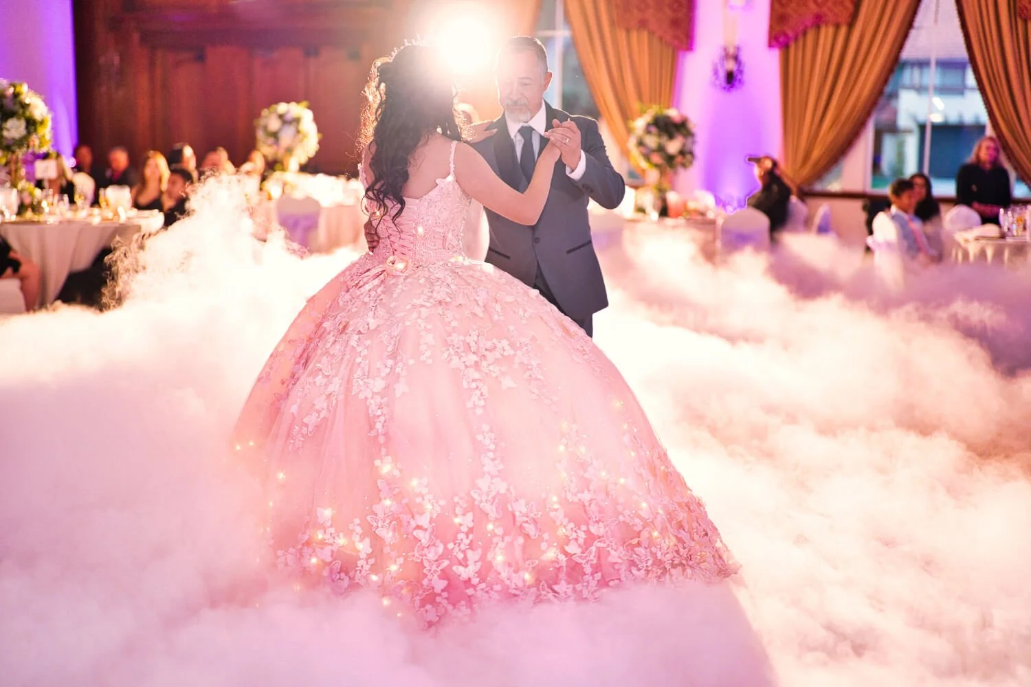 A bride and groom dance in a wedding reception with fog effects, surrounded by seated guests and decorated with flowers and curtains.