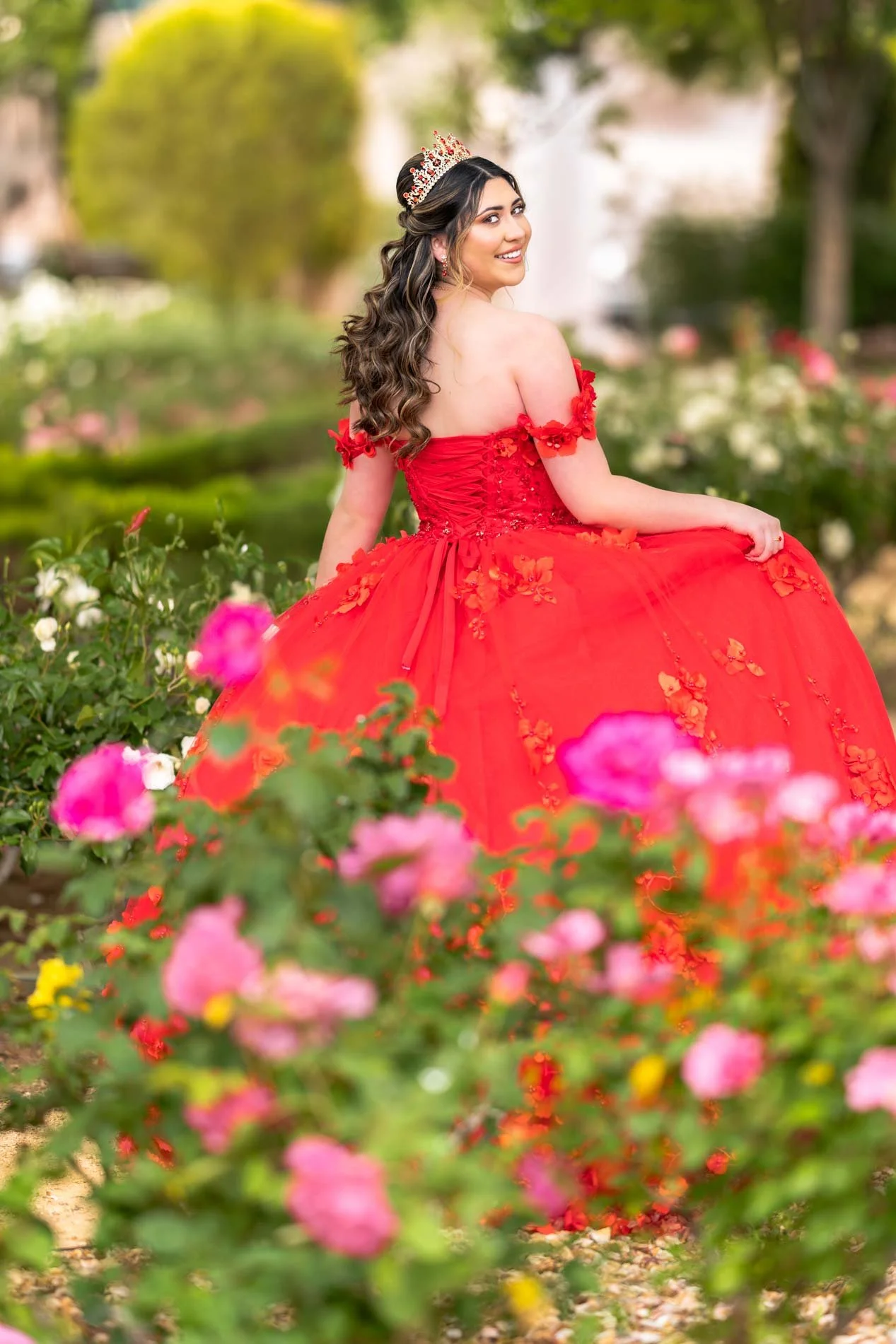Young woman in a red ball gown with floral details, wearing a tiara, sitting in a garden with pink and white roses, smiling over her shoulder.