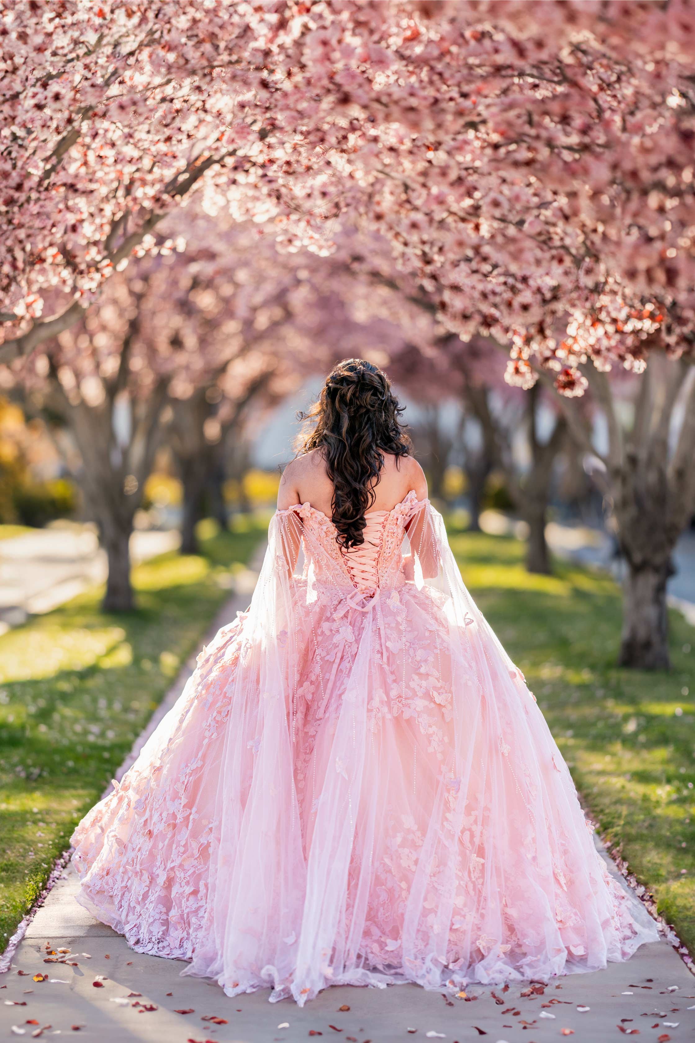 Woman in a light pink, elaborate ball gown with off-the-shoulder sleeves walking down a sidewalk lined with pink cherry blossom trees.