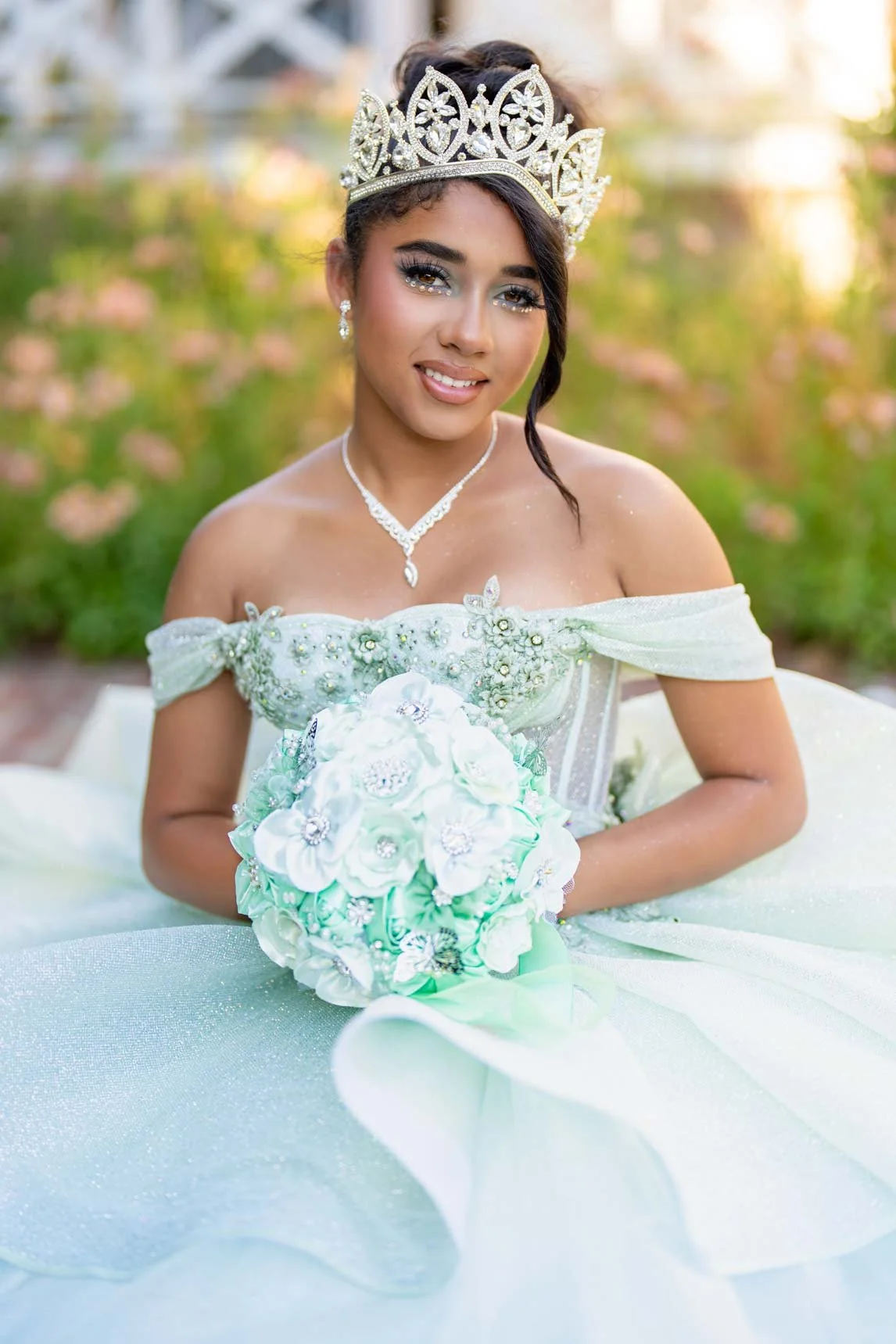 A young woman dressed in a white wedding gown with floral embellishments, wearing a tiara and jewelry, holding a bouquet, outdoors in a garden setting.