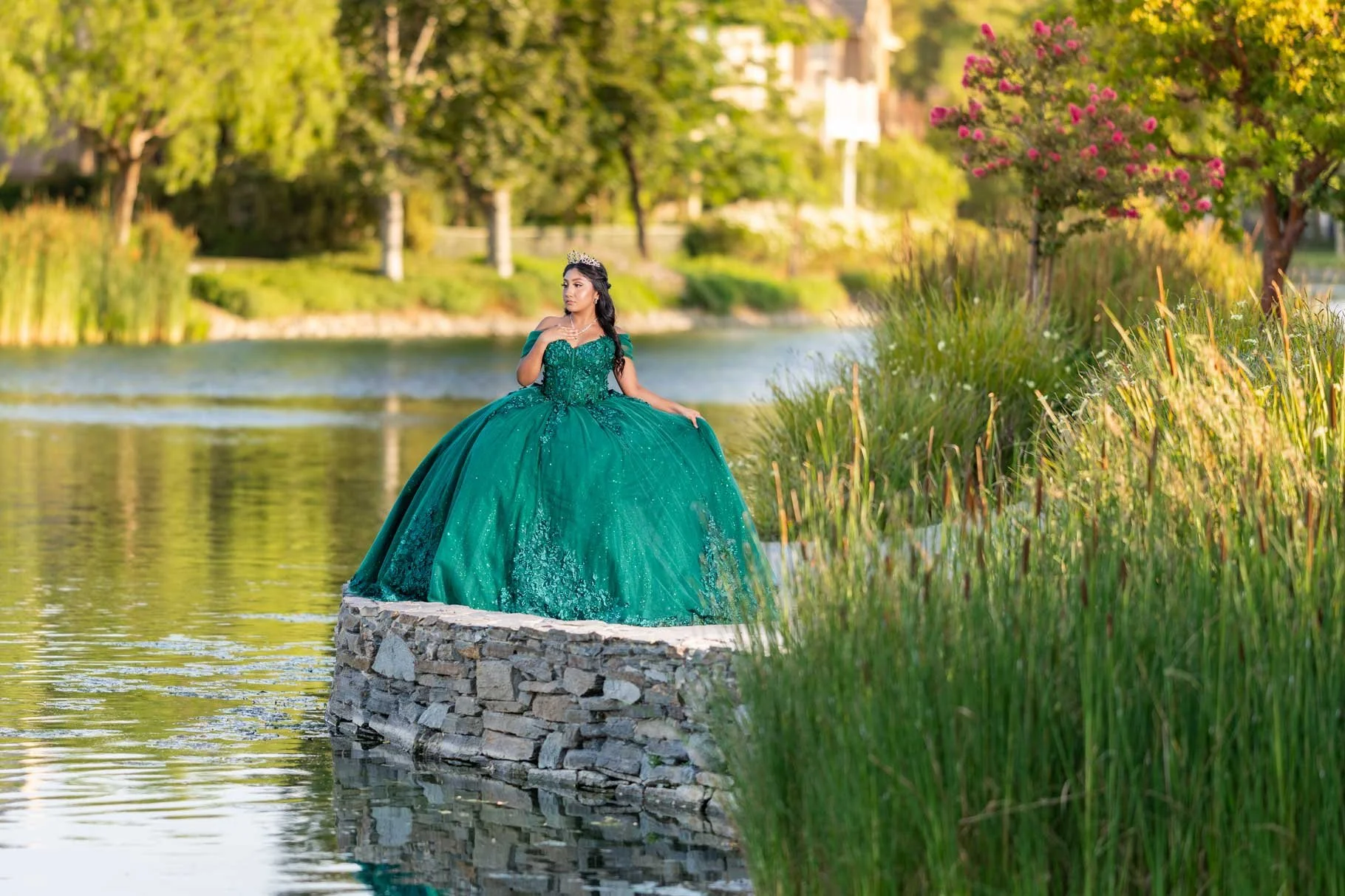 A woman in an emerald green ball gown and tiara standing on a stone platform by a river, with lush green trees and flowering bushes in the background.
