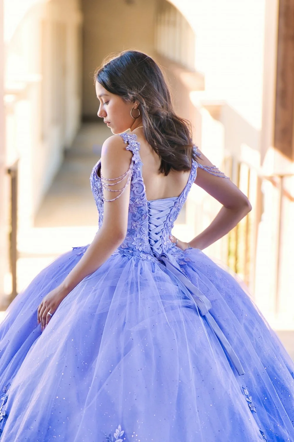 A woman in a lavender ball gown with lace and bead details, posing with her back to the camera, in a sunlit hallway.