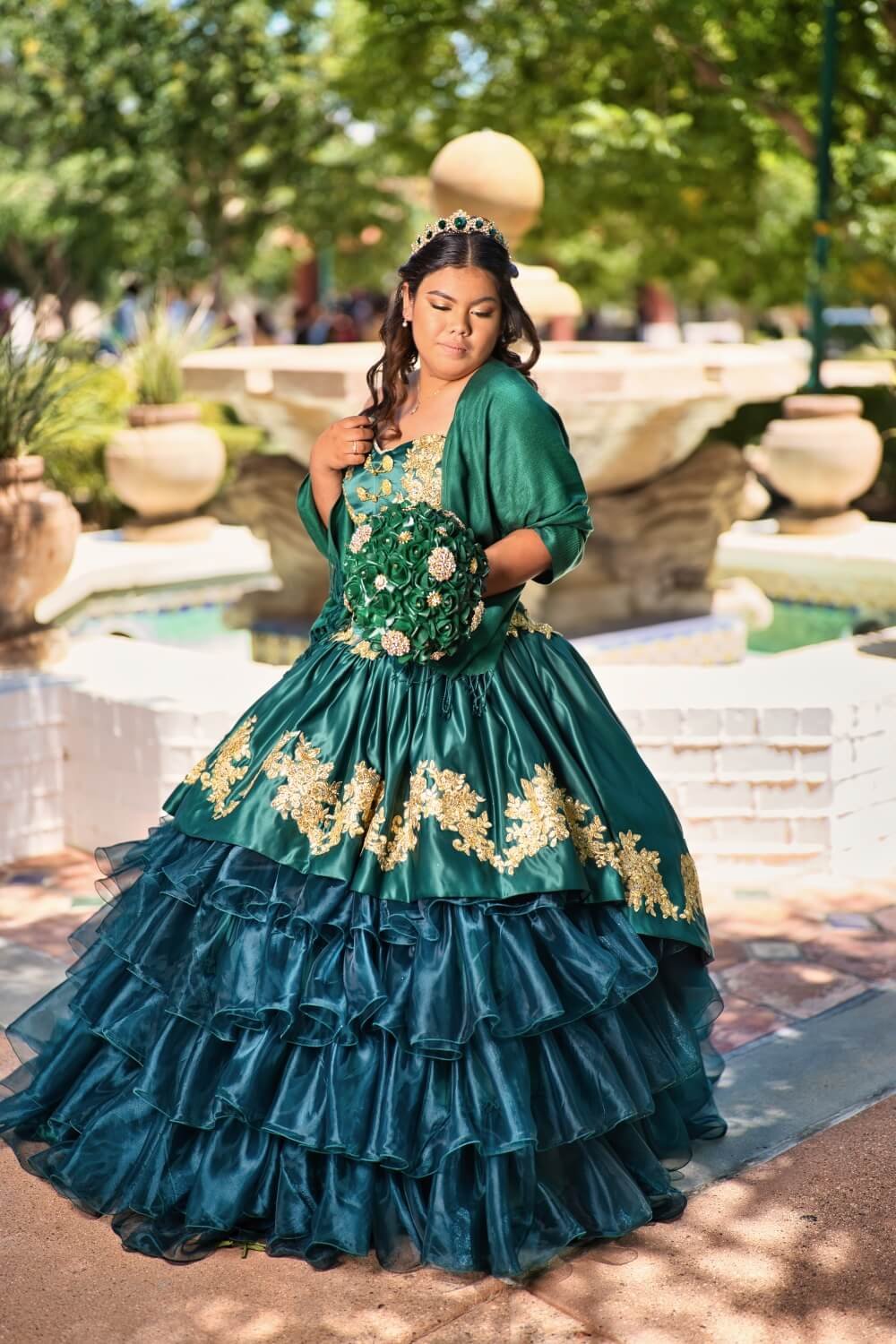 Young woman in a green and gold quinceañera dress holding a bouquet, standing outdoors near a fountain.